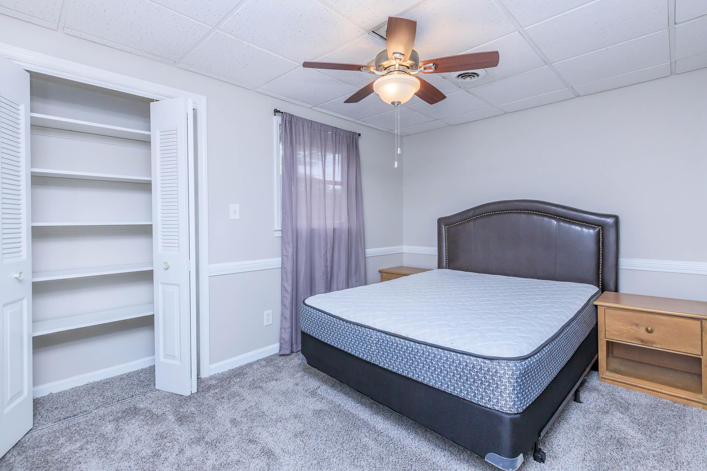 A cozy bedroom featuring a brown upholstered bed frame with a mattress, light gray walls, and a ceiling fan. There's a wooden nightstand beside the bed, and a closet with open shelves. A purple curtain hangs over a window, adding a touch of color to the neutral decor. Carpeted flooring enhances the room's warmth.