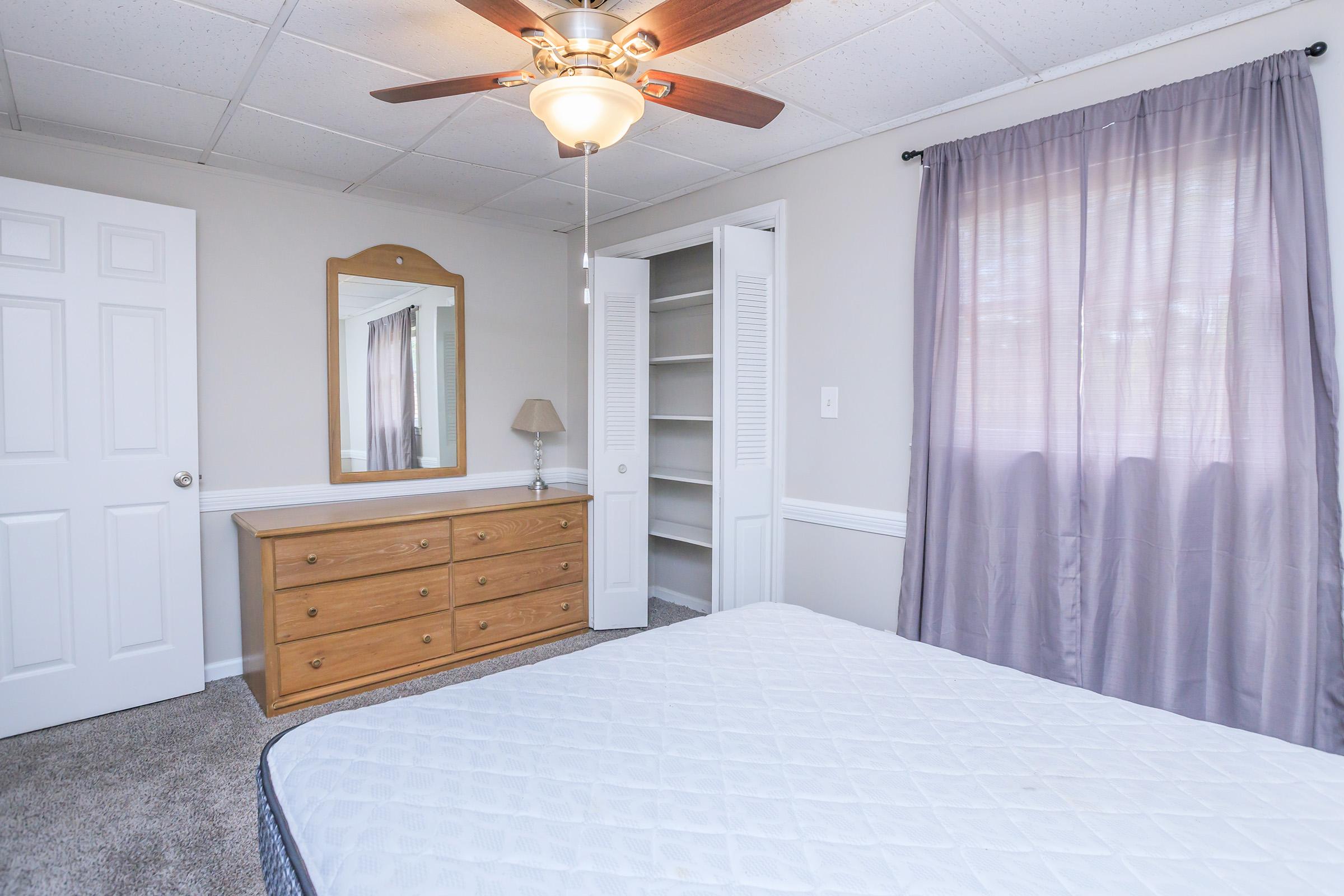 A well-lit bedroom featuring a light brown wooden dresser beneath a mirror, a ceiling fan, and a closet with double doors. The walls are painted a soft gray, and there are light purple curtains covering the window. The carpeted floor is neutral in color, and a bed with a white mattress is positioned in the room.