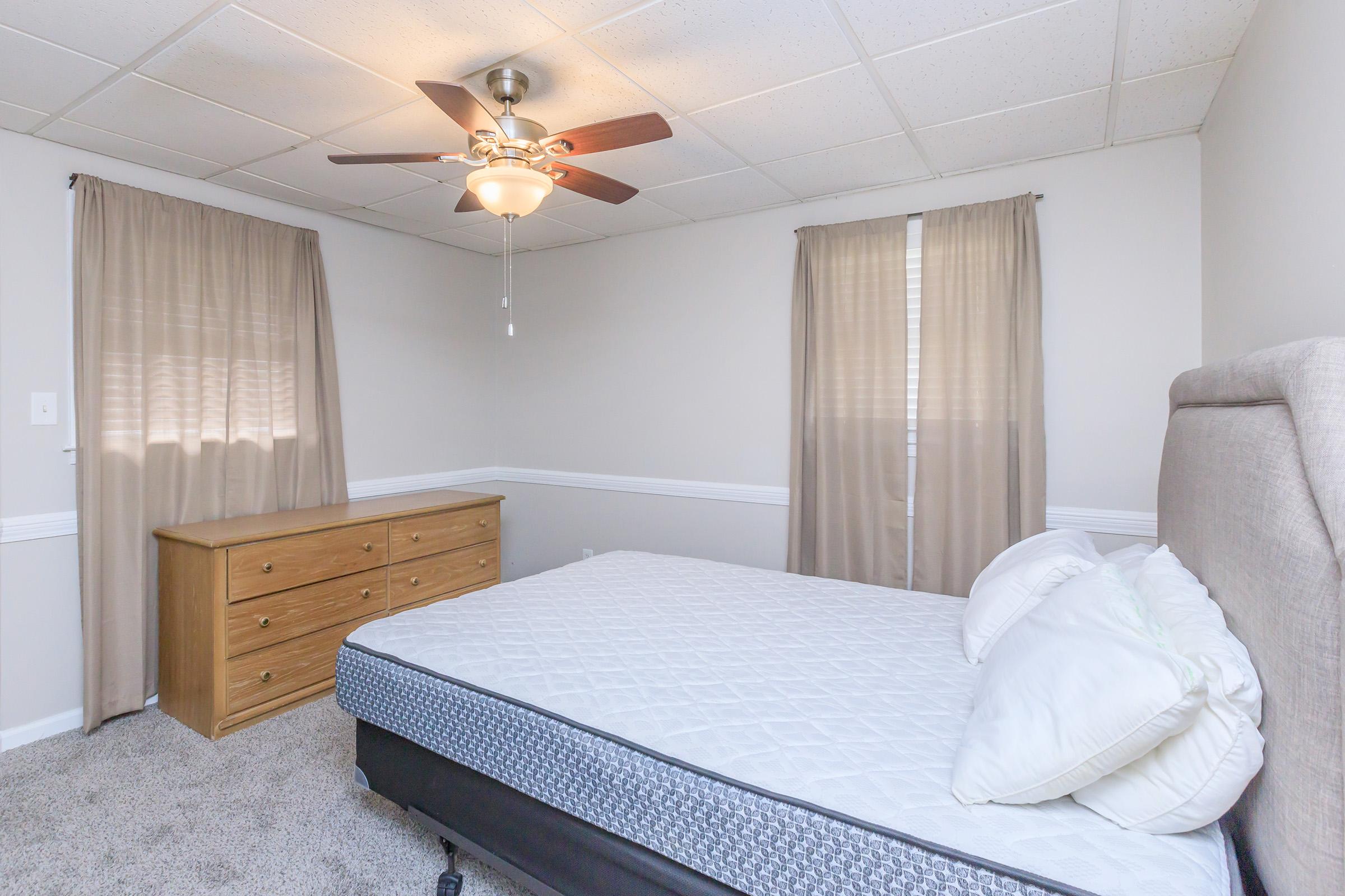 A well-lit bedroom featuring a queen-sized bed with a light gray upholstered headboard and white bedding. There are two curtains on the windows, a ceiling fan overhead, and a wooden dresser against the wall. The carpet is a soft gray, creating a cozy atmosphere.