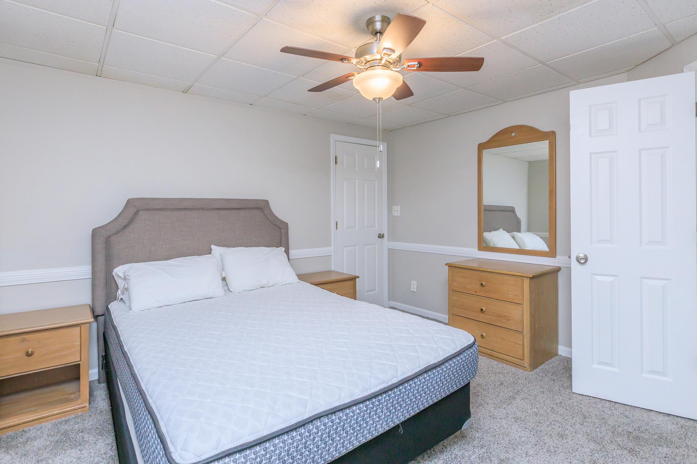 A tidy bedroom featuring a bed with a light gray headboard, white bedding, and two pillows. There's a wooden dresser with four drawers and a mirror near the bed, and a ceiling fan overhead. The walls are light-colored, and the floor is carpeted. A closed white door is visible in the background.