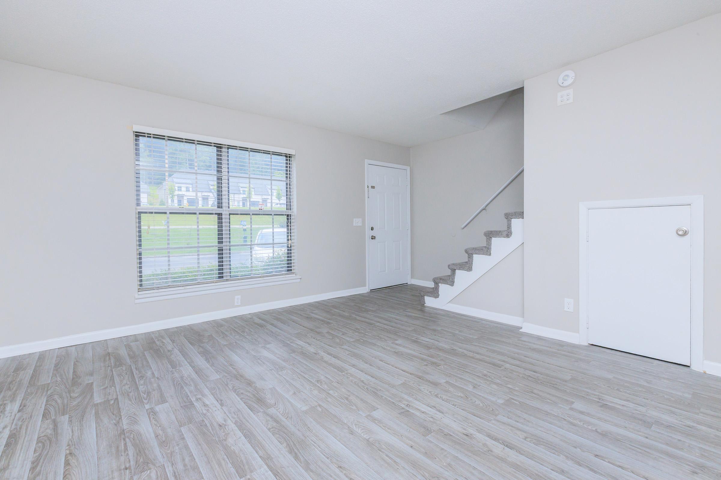 An empty living room with light gray walls and wood-style flooring. A large window with blinds allows natural light, and there is a staircase leading to the upper level. A door leads outside, and a small closet or storage space is visible under the stairs.