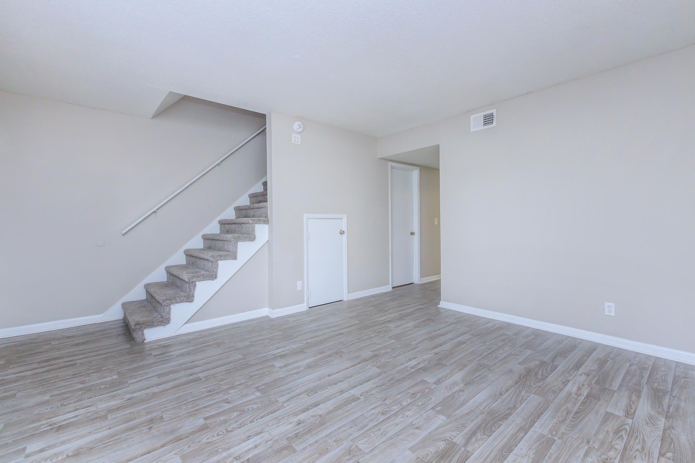A spacious, light-colored living area featuring a staircase with beige carpeting and wooden-style flooring. The walls are painted in a neutral tone, and there is a small door under the staircase, with an open doorway leading to another room. The overall ambiance is bright and modern.