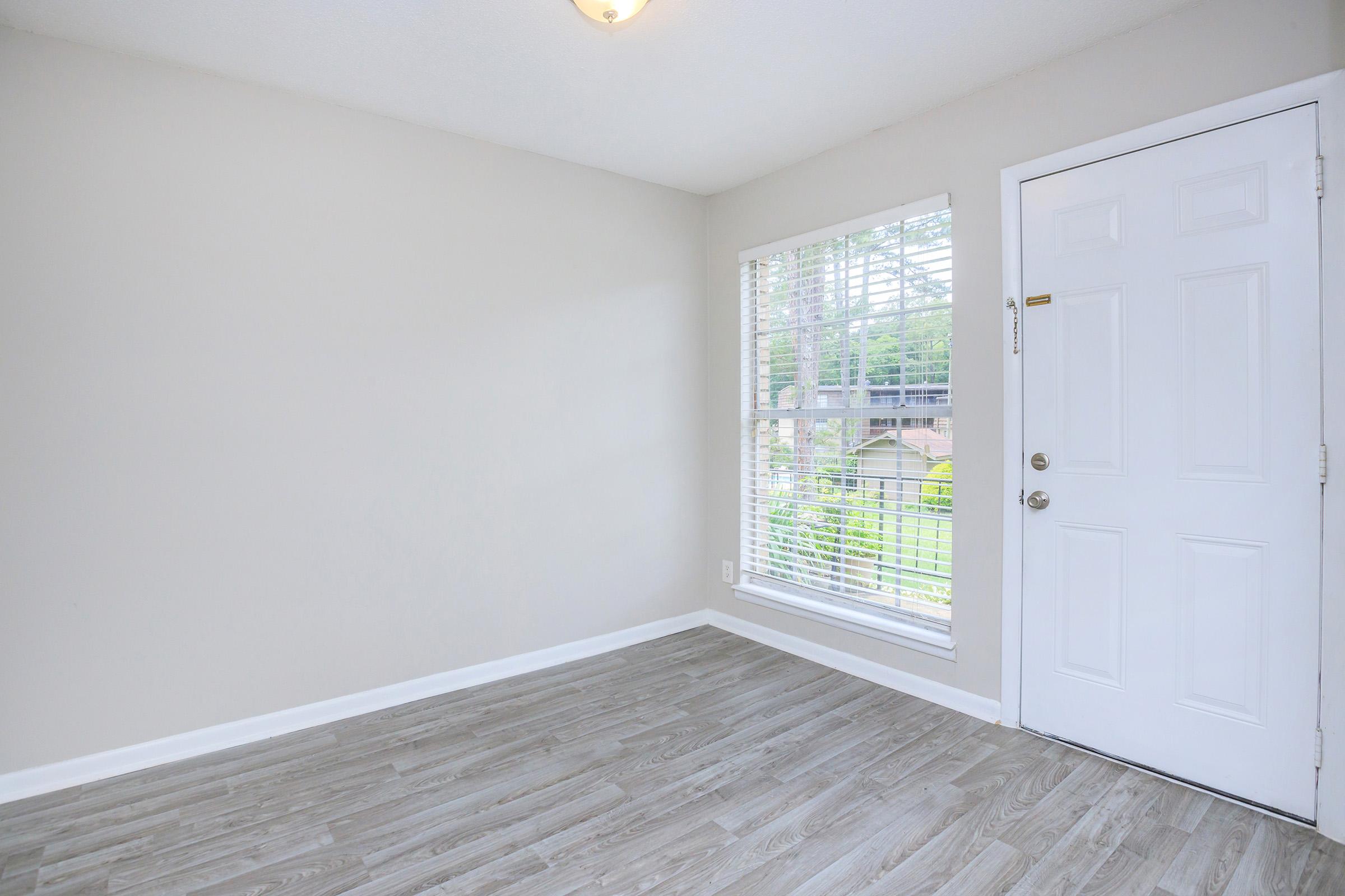 A clean, empty room with light gray walls and brown vinyl flooring. A large window with white blinds allows natural light in, and a white door is visible on the right side. The room has a bright and open feel, designed for potential furnishing or decoration.