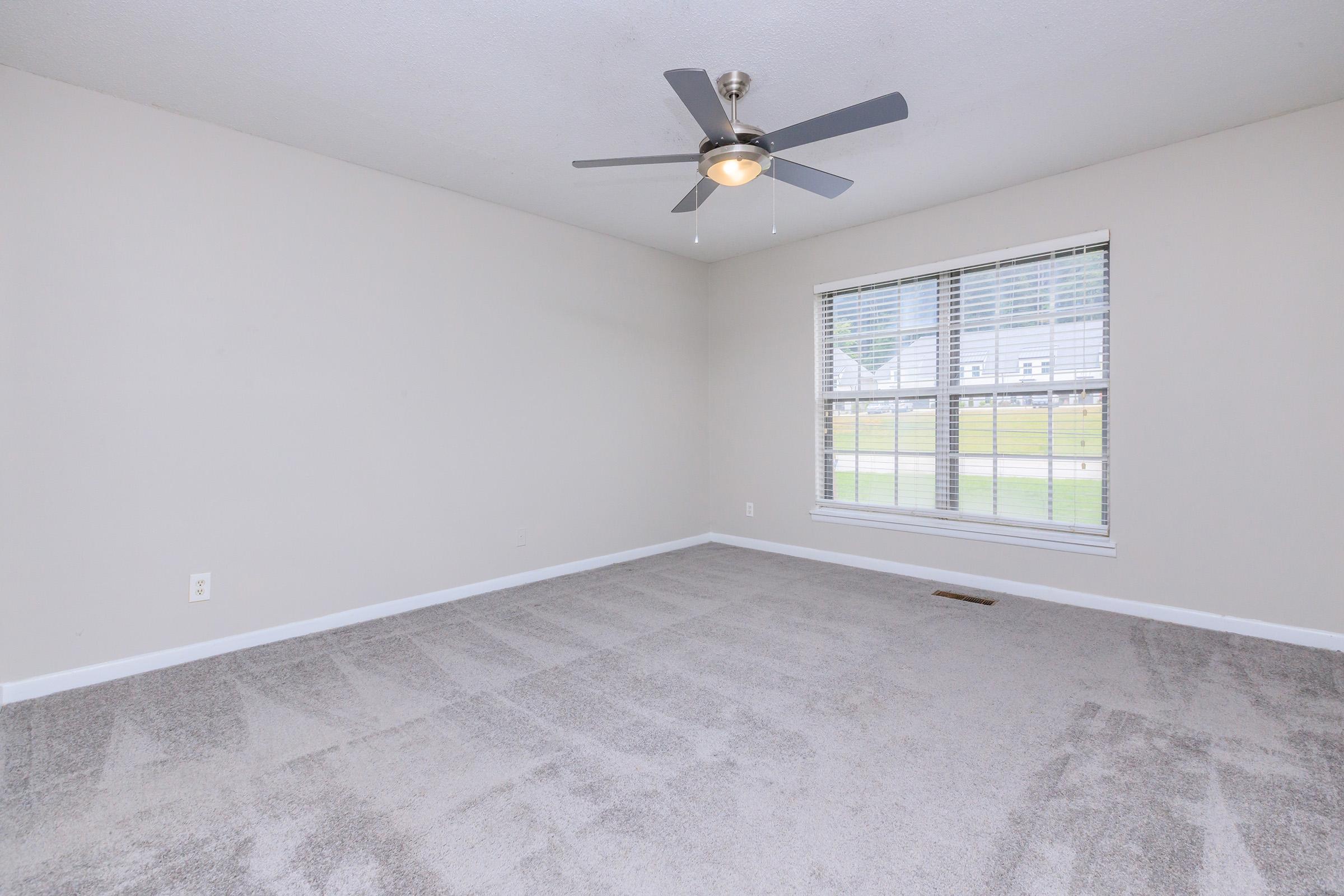 A spacious, empty room with light gray carpet and walls. The room features a ceiling fan with a light fixture and a large window with closed blinds, allowing natural light to enter. The space is uncluttered and ready for furnishings, creating a neutral and airy atmosphere.