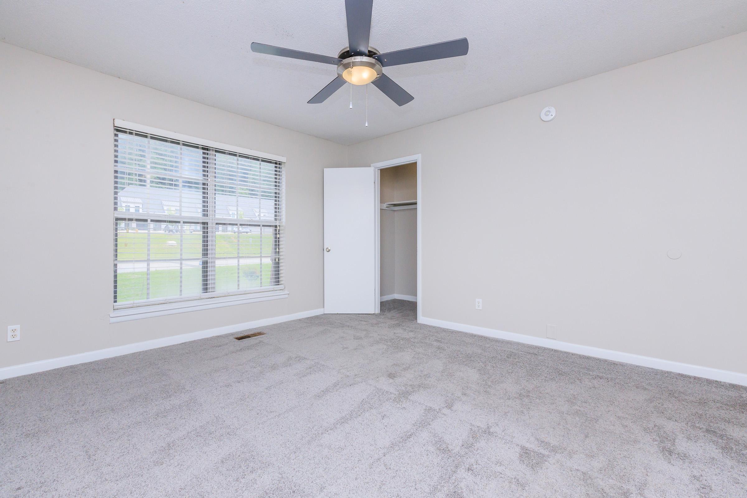 A spacious, empty bedroom featuring light beige walls, a ceiling fan, and a large window with horizontal blinds. The room has freshly installed gray carpet and a closet with an open door. Natural light streams in through the window, creating a bright and inviting atmosphere.