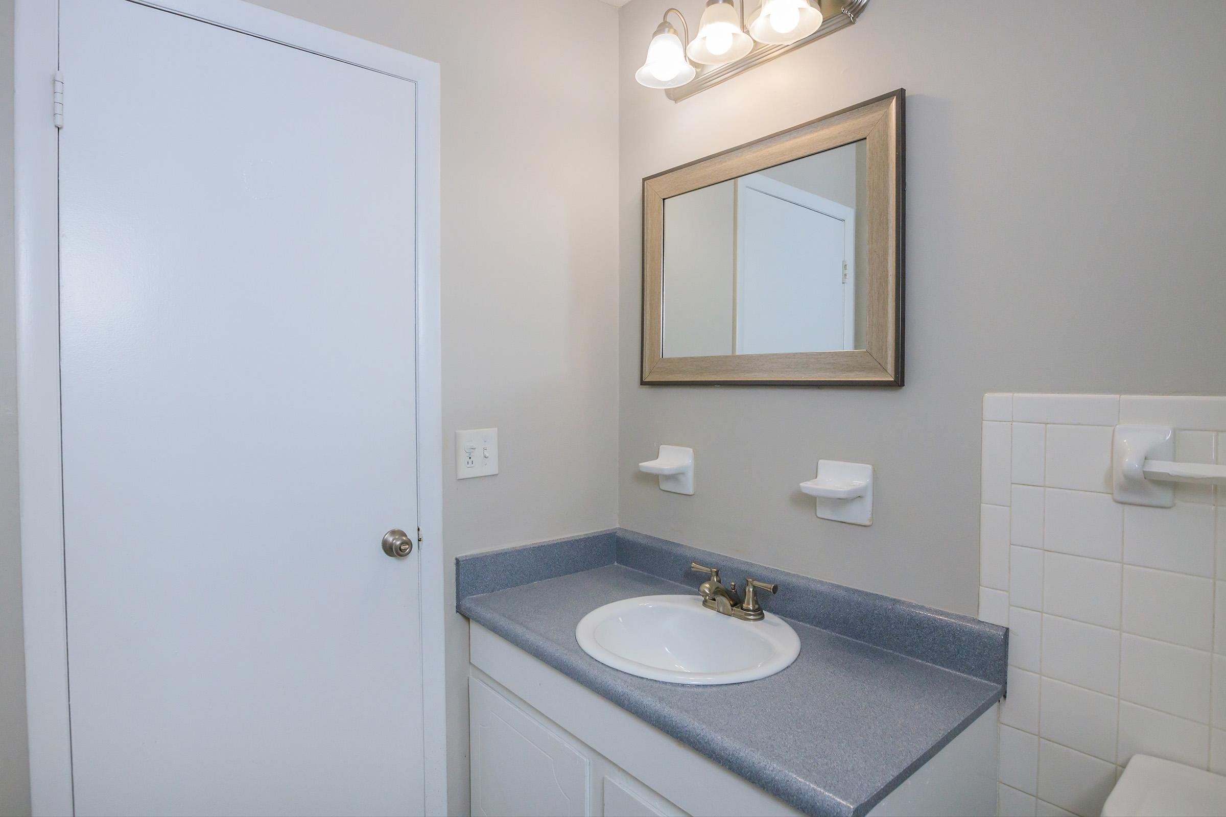 A simple bathroom interior featuring a white sink with a gold faucet, a light gray wall, a mirror with a decorative frame above the sink, two white soap holders mounted on the wall, and a closed door. The counter is blue, and there's a white tiled section with a towel rack nearby.