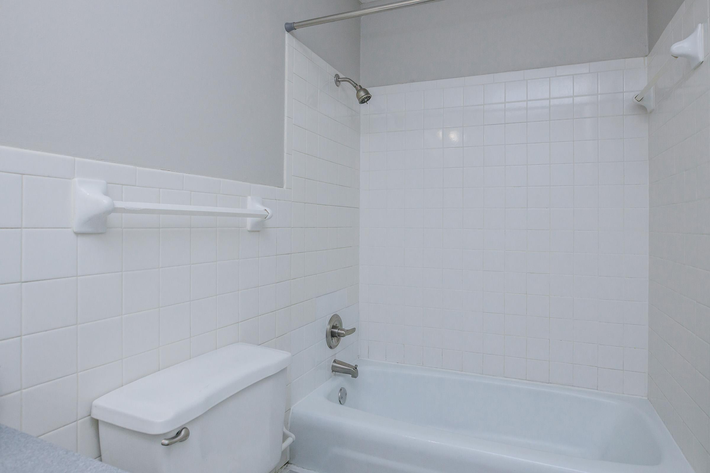 A clean bathroom featuring a white tiled shower area with a bathtub, a chrome faucet, and a toilet. The walls are painted in light gray, and there are grab bars installed on two sides of the shower for safety. Natural light brightens the space through an unobtrusive opening above.