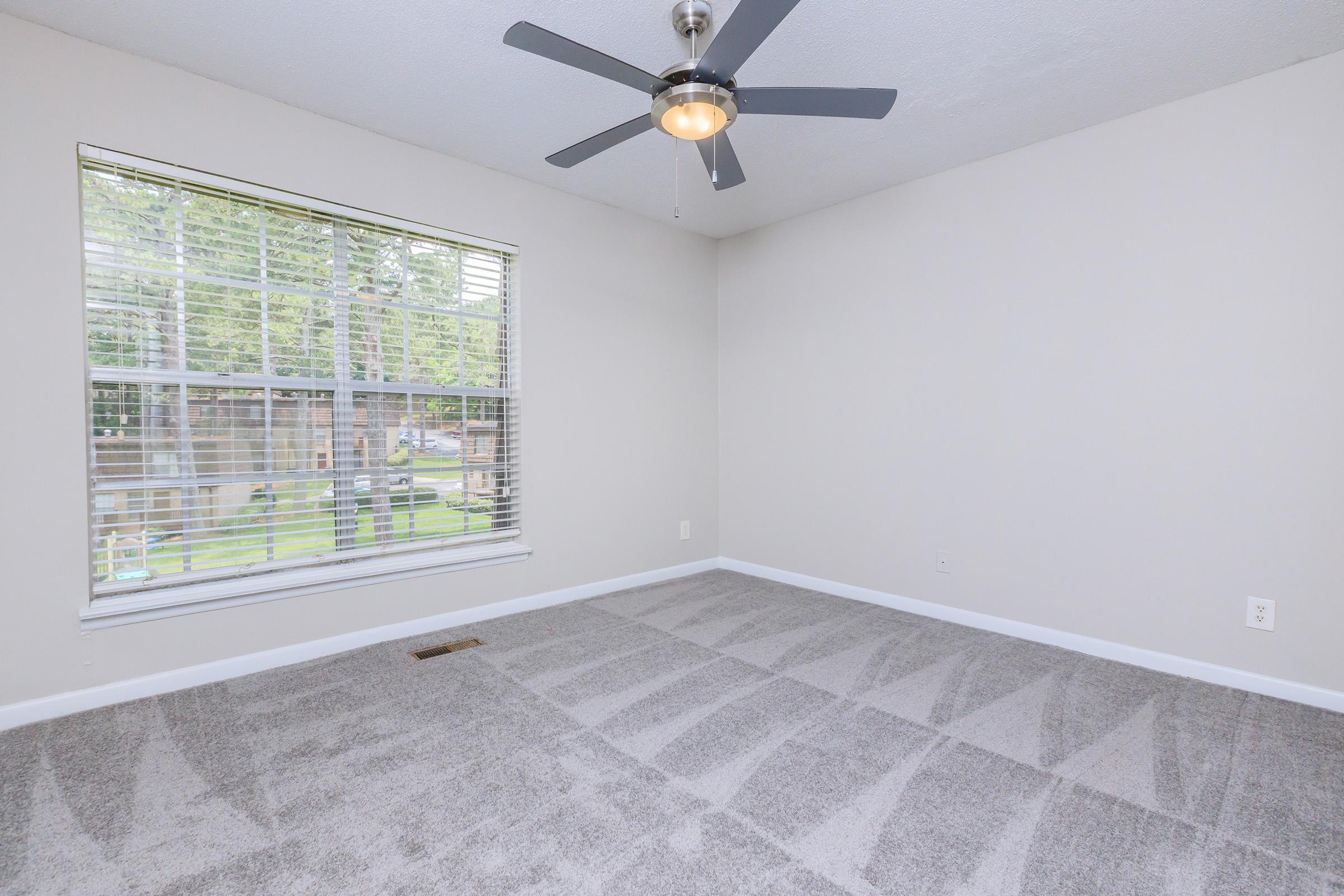 A vacant room with gray carpet and light gray walls. A large window with blinds allows natural light to enter, and a ceiling fan is installed. The space is clean and uncluttered, suggesting it is ready for new furnishings or decoration.