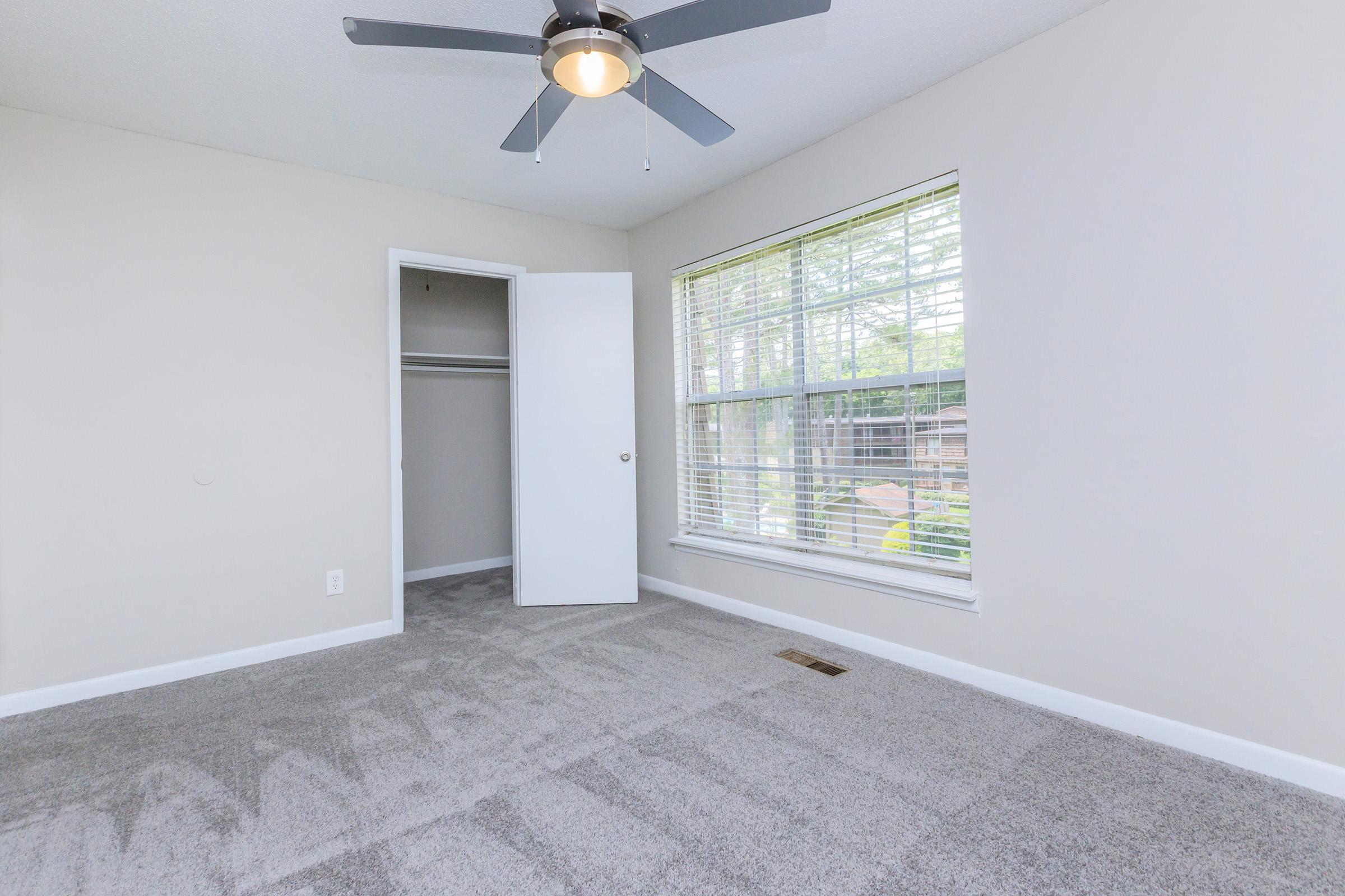 A bright, empty room featuring gray carpet, a ceiling fan, and a large window with blinds. There is an open closet with sliding doors on one side. The walls are painted a light color, creating a spacious and airy feel.