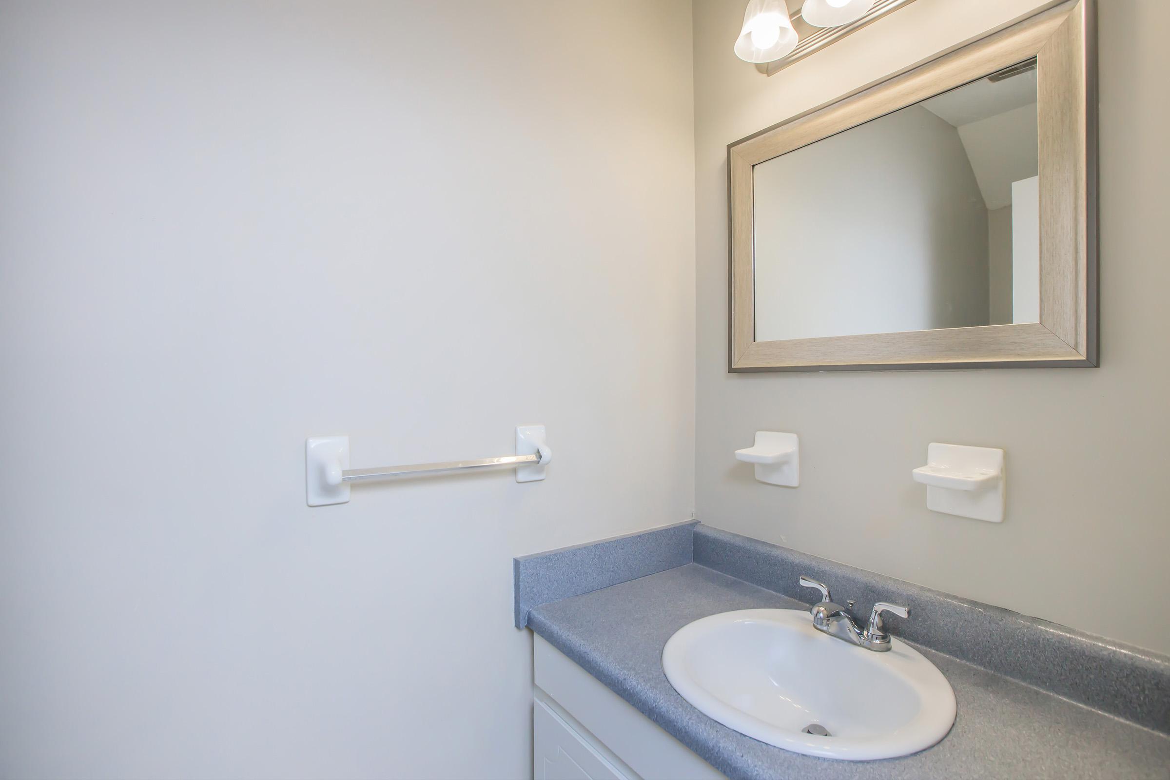 A clean and modern bathroom with a light gray wall, a sink with a faucet, and a mirror above. The counter is light blue with two soap dishes mounted on the wall and a towel rack beside the sink. Soft lighting from two fixtures above the mirror creates a bright atmosphere.