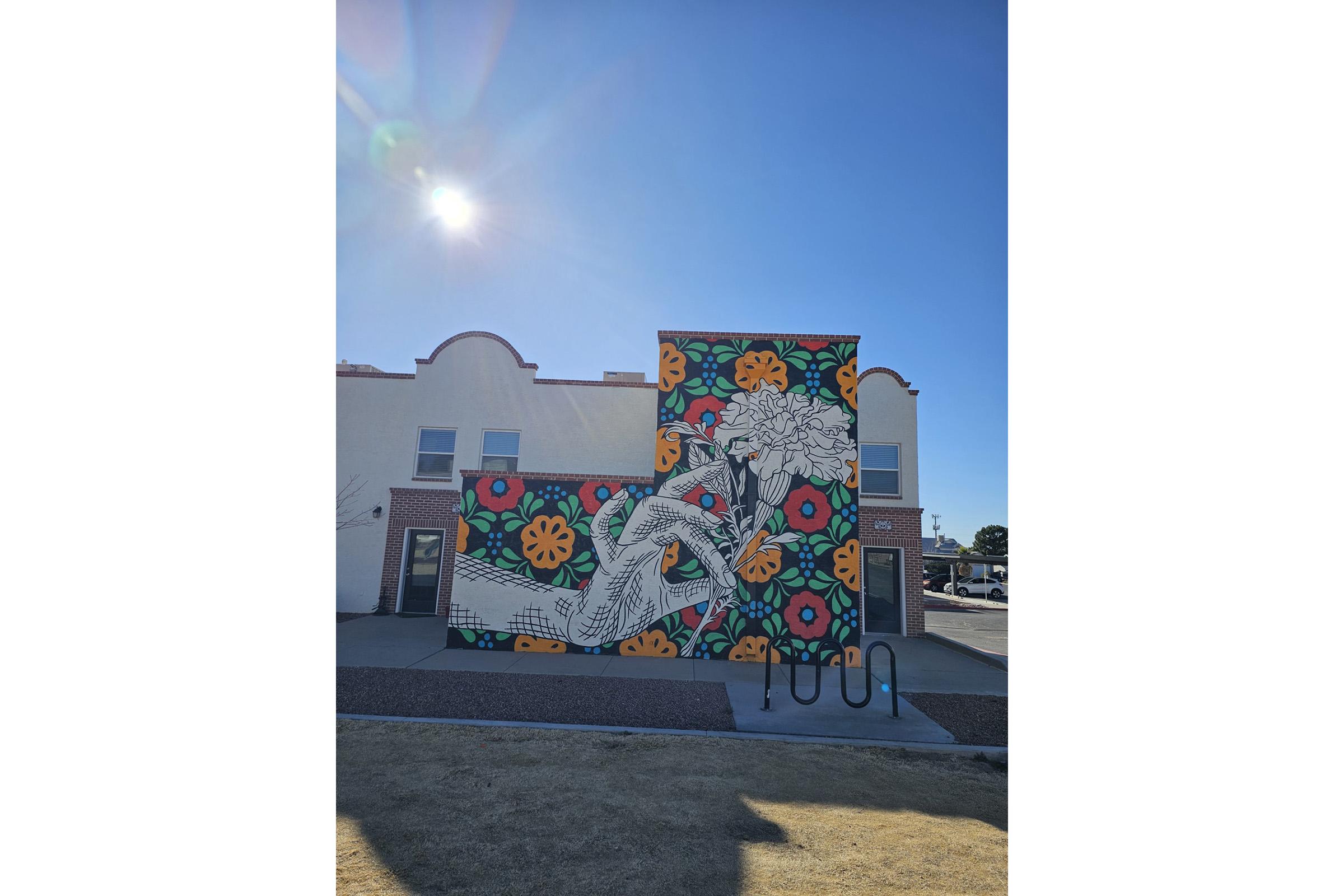 A vibrant mural on a building featuring a hand drawing flowers amidst colorful floral patterns. The background consists of diverse bright colors, including orange, pink, and green, creating a lively and artistic atmosphere. Sunny skies are visible above, adding to the cheerful scene.