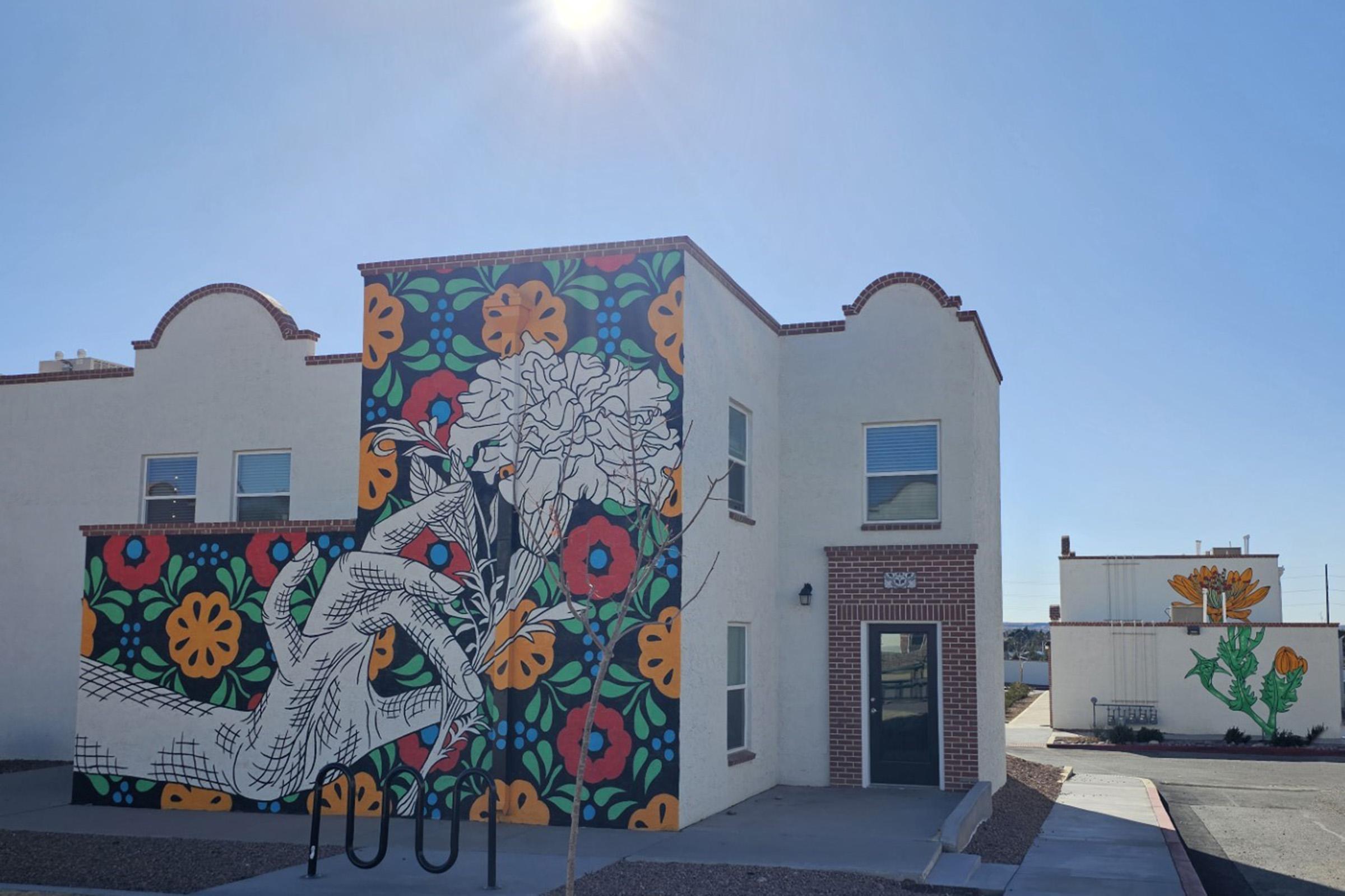 A vibrant mural on the side of a building features a hand holding a flower, surrounded by colorful floral patterns in red, yellow, and green. The building is two stories tall with a white exterior and a door on the front. Sunlight reflects off the structure, creating a bright atmosphere.