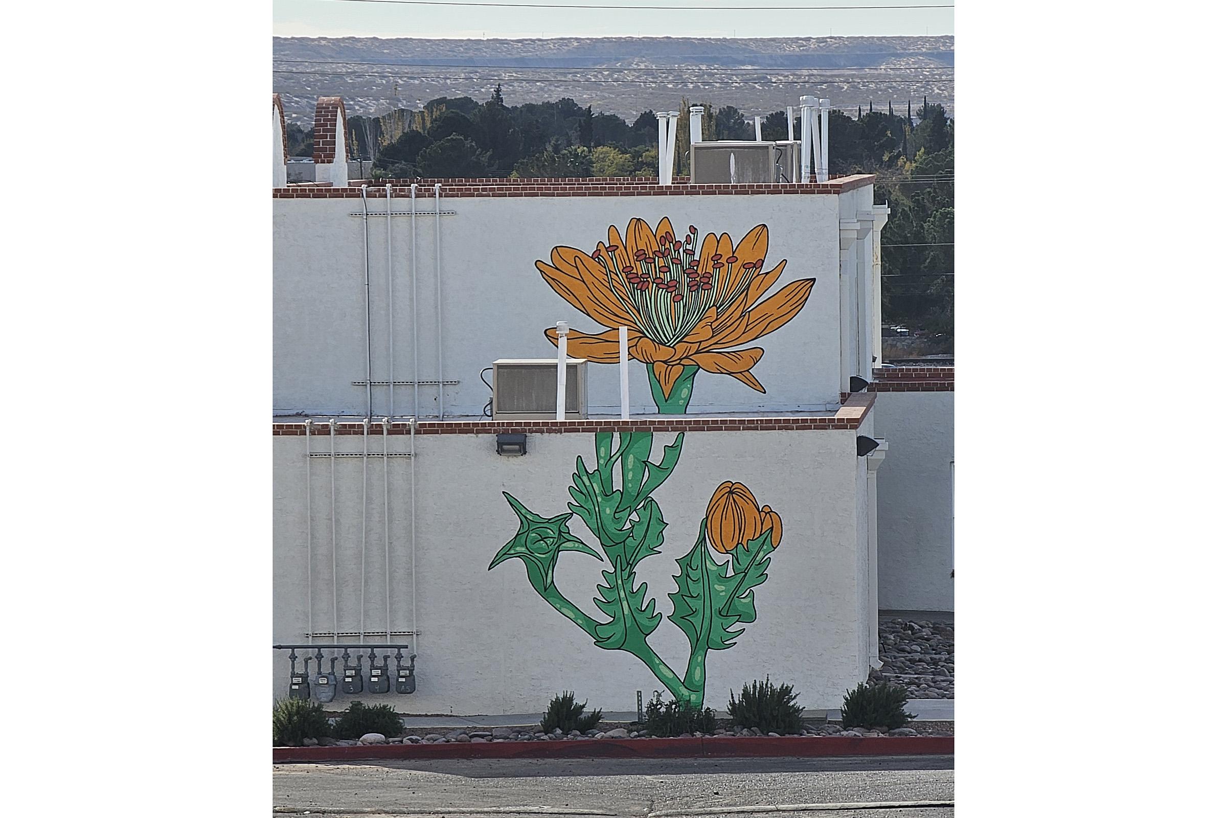 A vibrant mural depicting a large orange flower with green leaves and stem, painted on the side of a white building. The artwork features detailed line work, with the flower prominently displayed, adding color and character to the urban landscape.