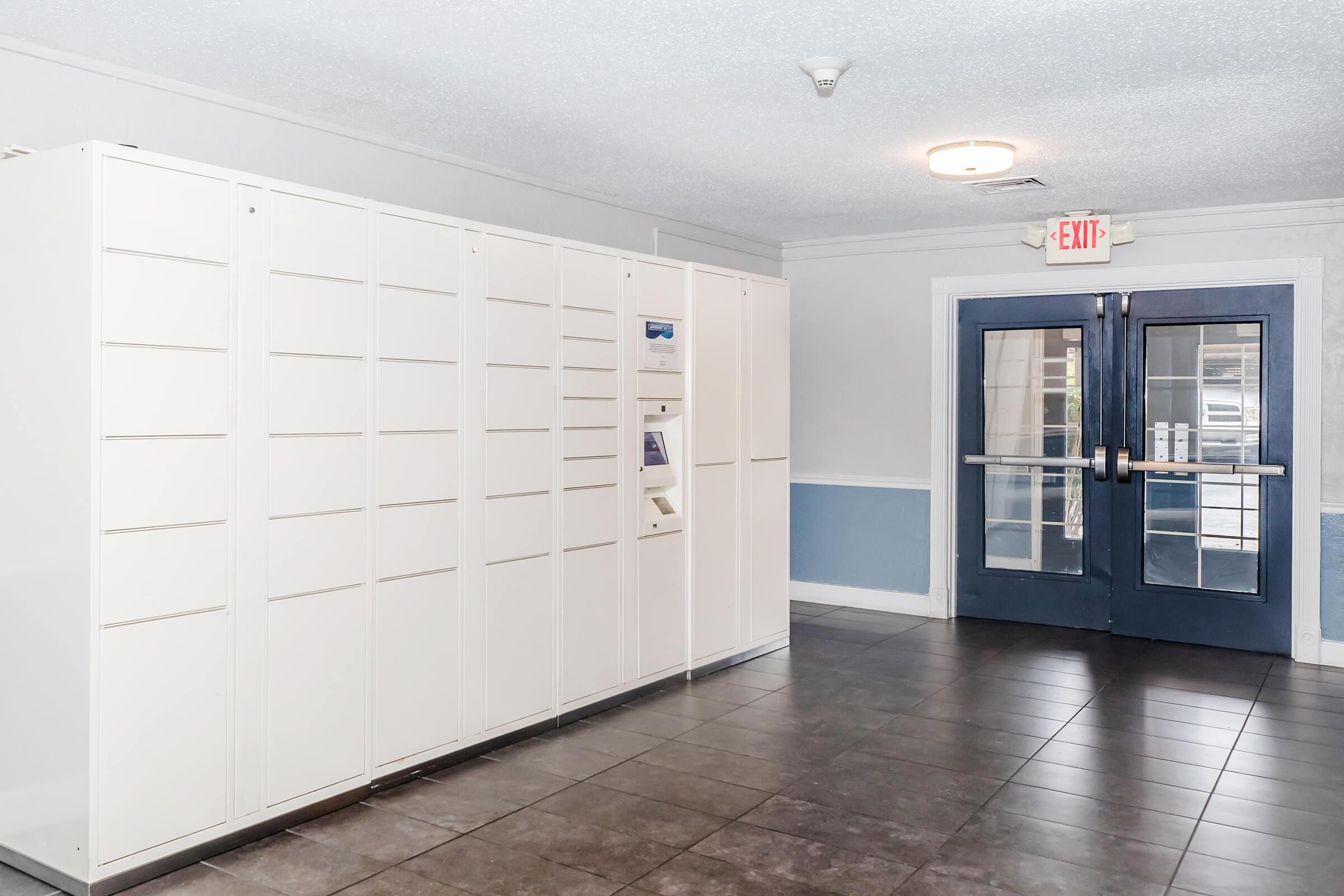 A modern hallway featuring a white locker system for package delivery alongside double glass doors leading outside, with a light-colored wall and dark tiled floor. The environment appears clean and well-lit.