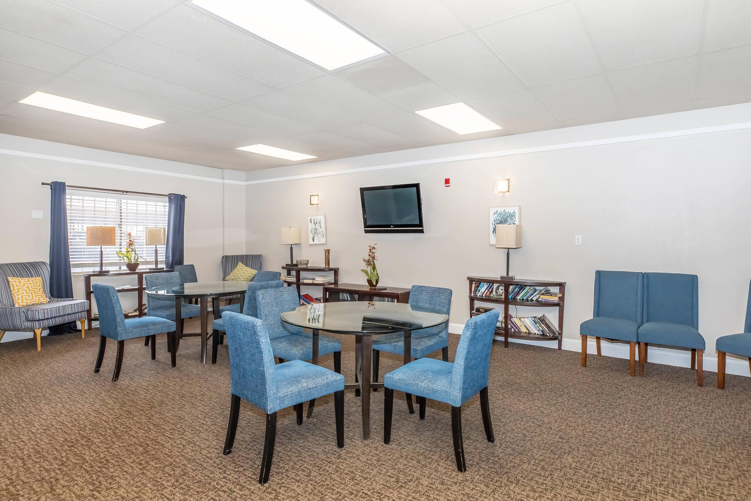 A well-lit common area featuring several round tables with blue chairs, a cozy seating area with an armchair, and a television mounted on the wall. Shelves with books are visible, and windows provide natural light, enhancing the welcoming atmosphere.