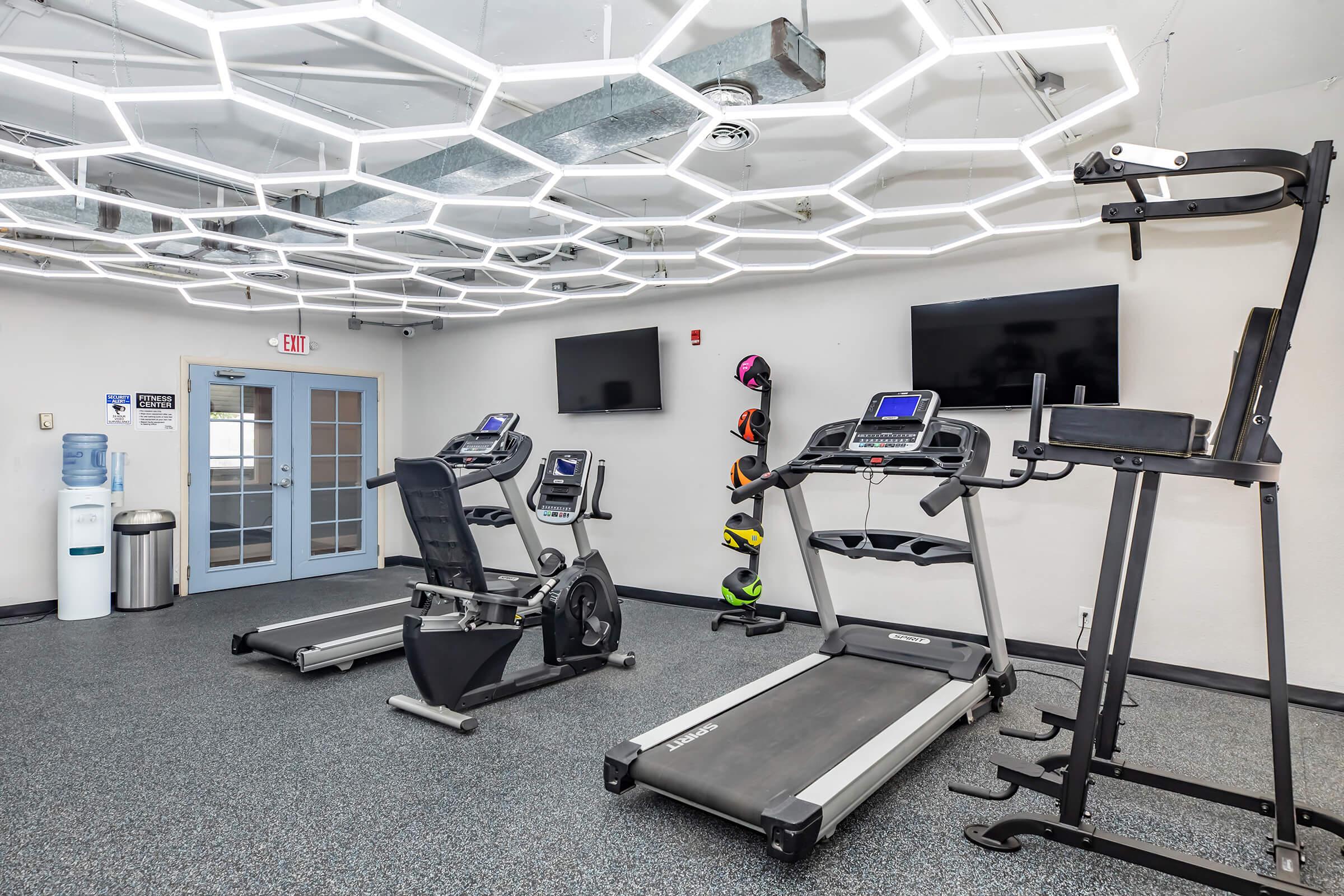A modern gym interior featuring exercise equipment including a treadmill, stationary bike, and an elliptical machine. The room is well-lit with a unique geometric ceiling light design, and there are two wall-mounted TVs. A water cooler is visible in the corner, along with colorful helmets displayed on the wall.