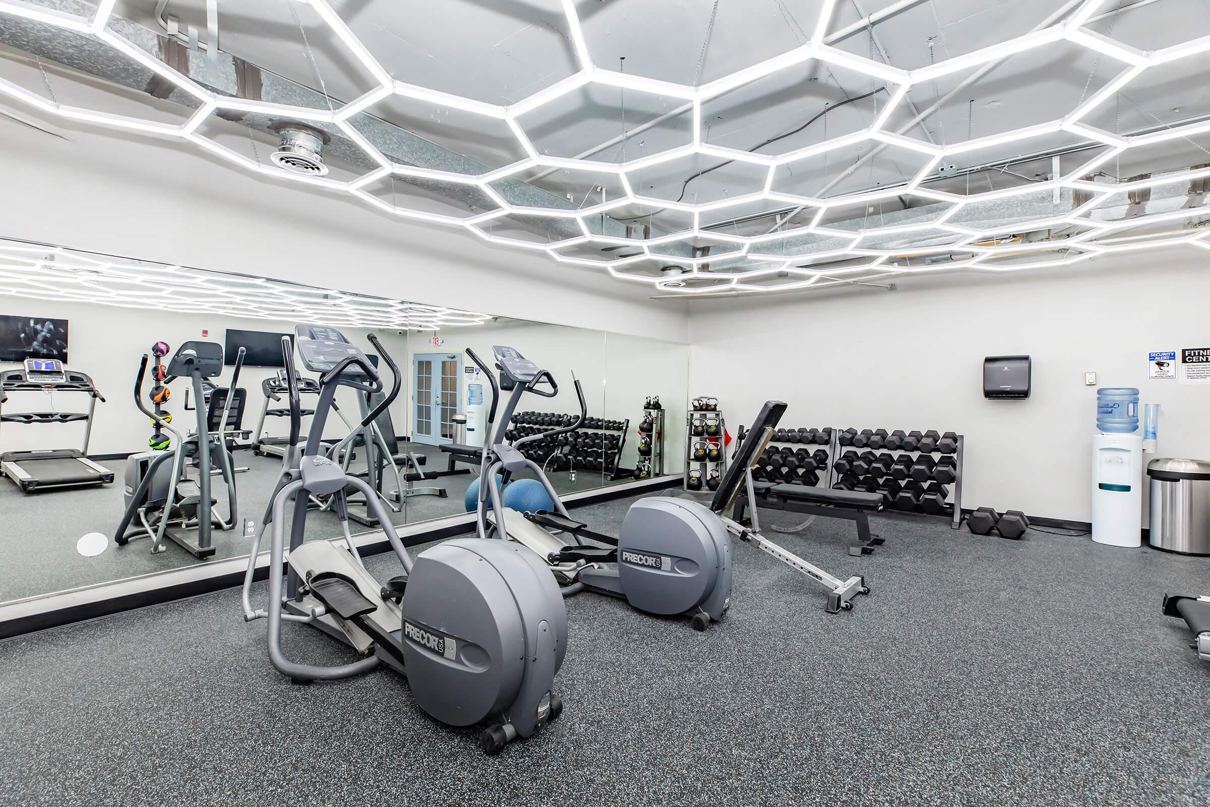 A modern gym interior featuring elliptical machines and a rowing machine. There are free weights neatly organized on a rack in the background. The ceiling has a distinctive hexagonal light design, adding a contemporary feel to the space. A mirrored wall reflects the workout equipment.