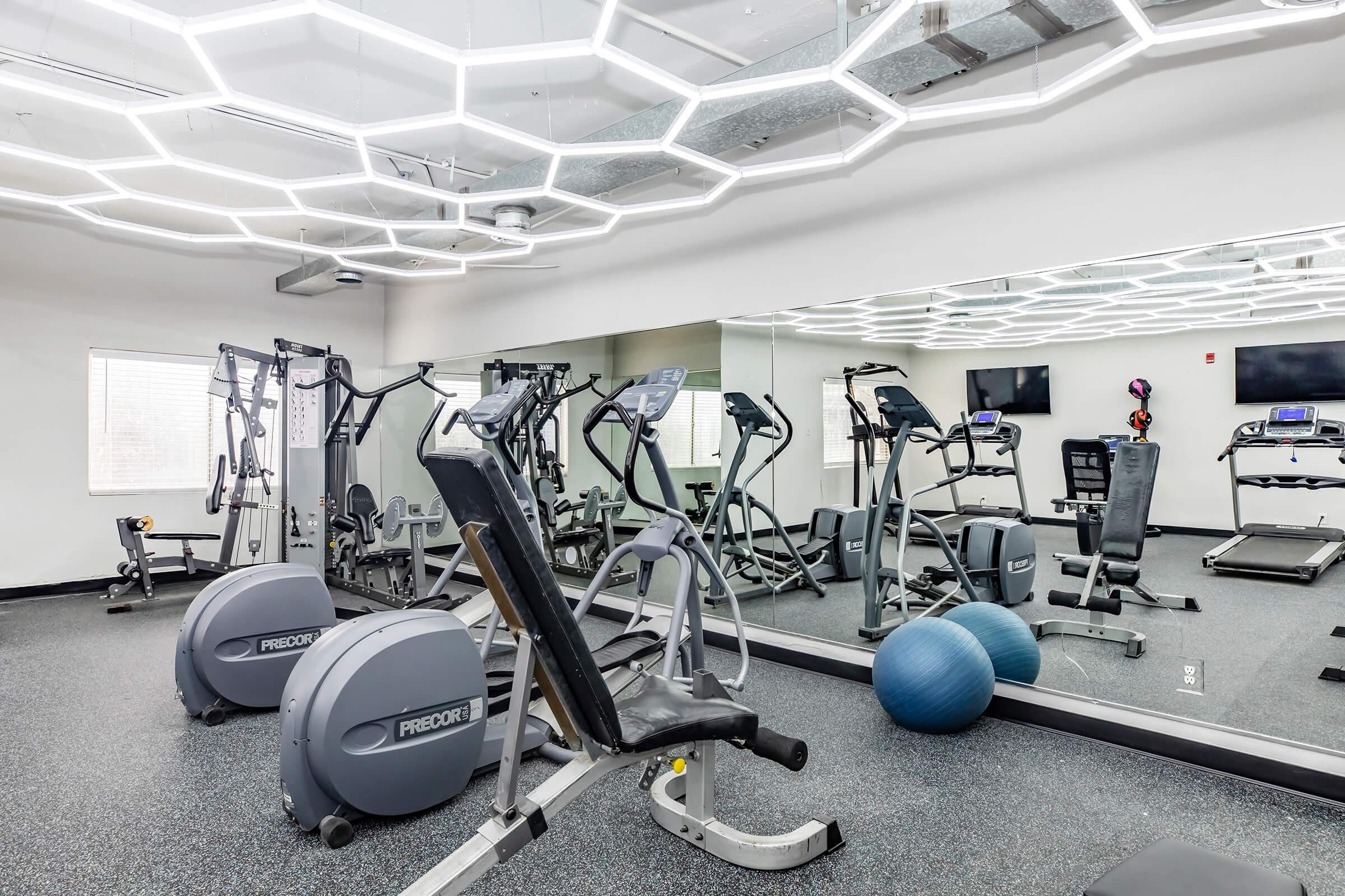 Bright and modern gym interior featuring various workout equipment, including ellipticals, a weight machine, and treadmills. The ceiling has a unique hexagonal light design, and there are mirrors on one wall along with exercise mats and a stability ball on the floor.