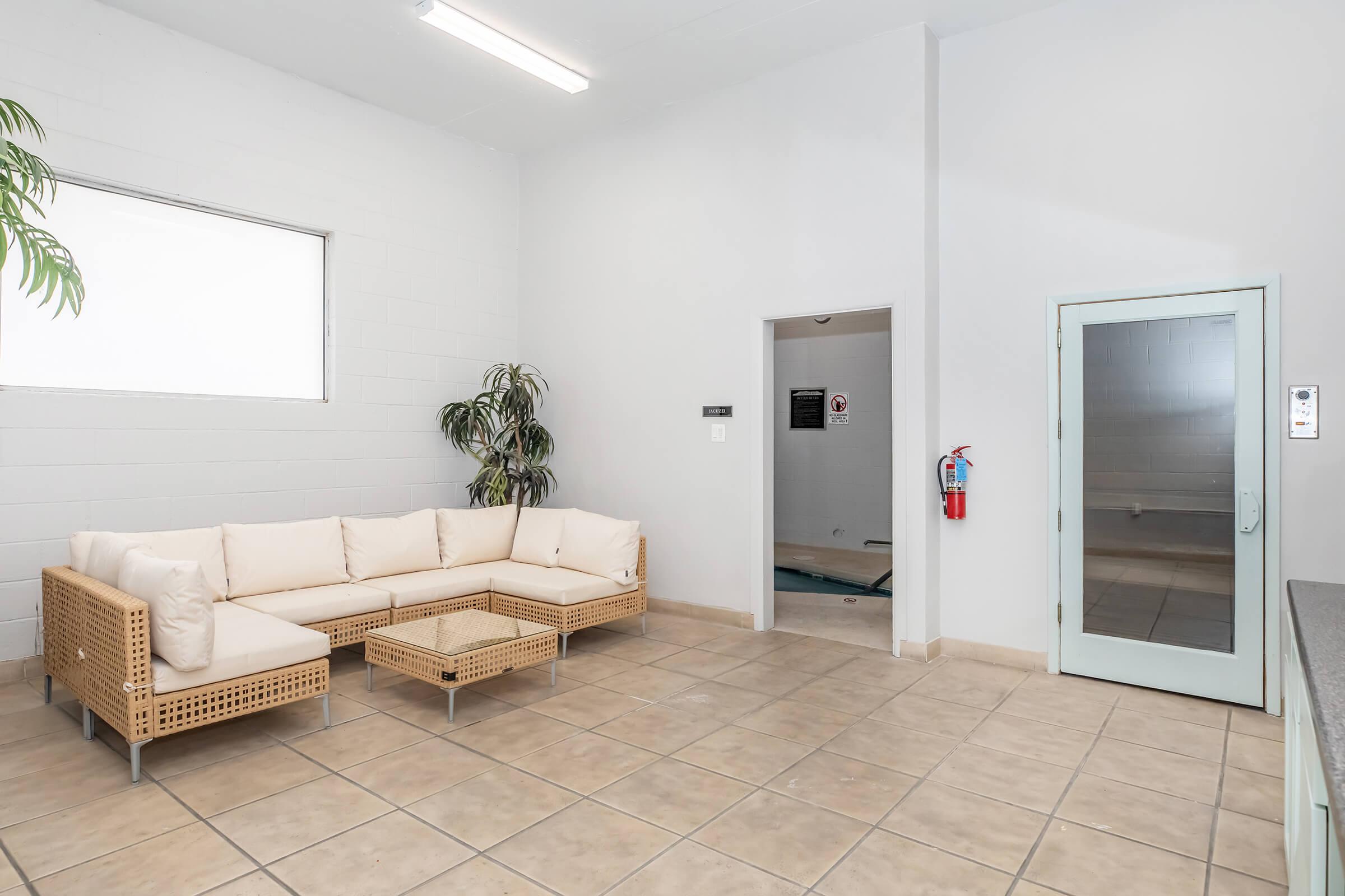 A bright room with a light-colored tiled floor, featuring a large, cream-colored sectional sofa with a glass coffee table. A potted plant adds a touch of greenery. There is a doorway leading to another room, and an exterior door with a push button for entry. The walls are plain and white, enhancing the spacious feel.