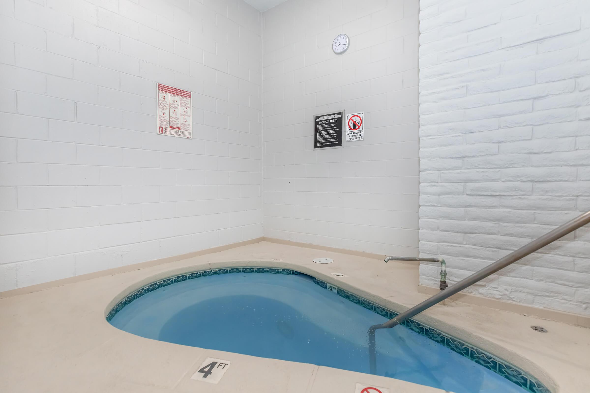 A small indoor hot tub with clear water, surrounded by white walls. There are safety signs posted nearby. A clock is mounted on the wall, and the water depth marker reads "4 FT." A metal handrail leads into the tub area.