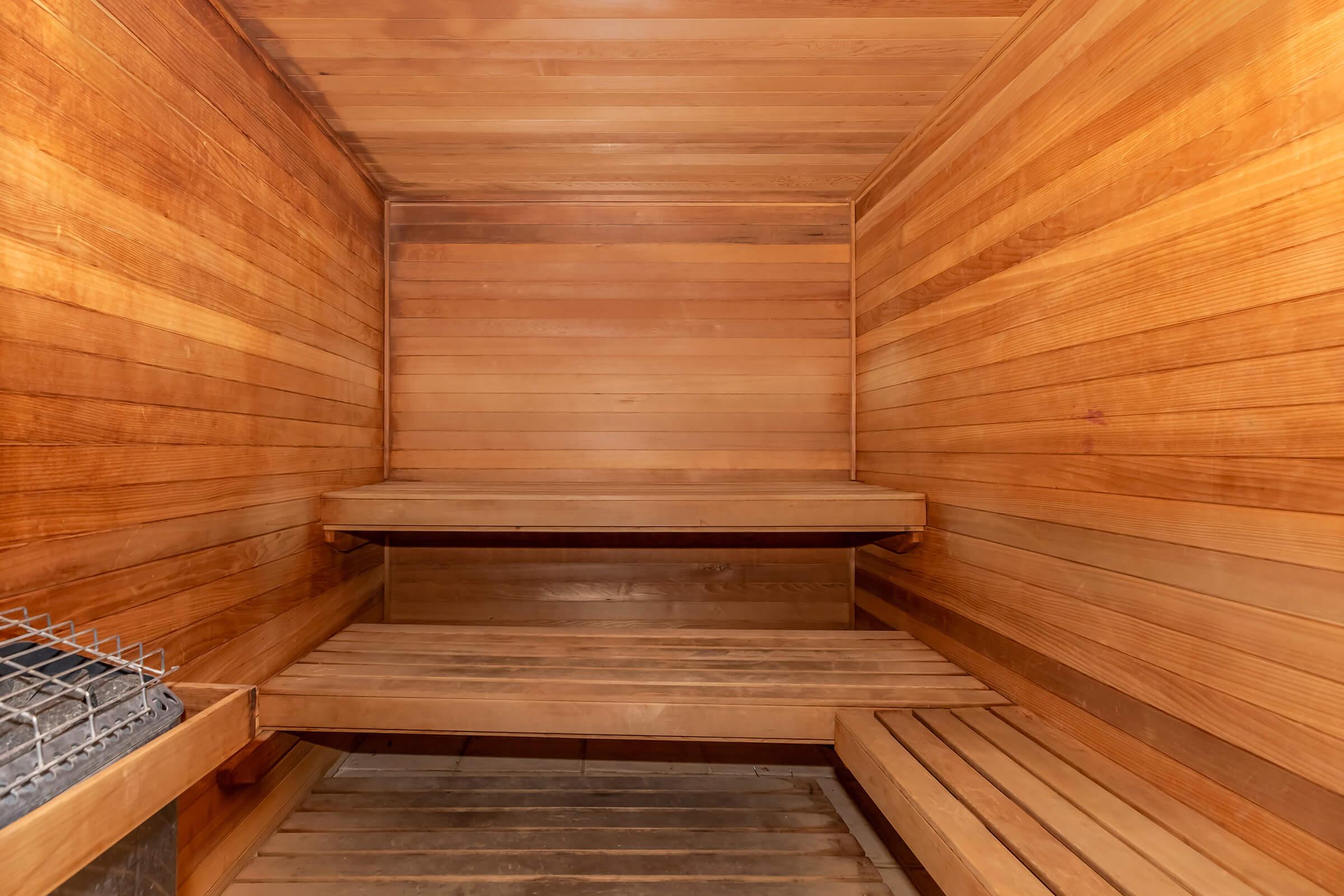 Interior of a wooden sauna featuring smooth, polished wooden walls and benches. The design is simple and rustic, with a stone heater located in one corner. The atmosphere is warm and inviting, ideal for relaxation and wellness. The lighting is soft, enhancing the serene ambiance.