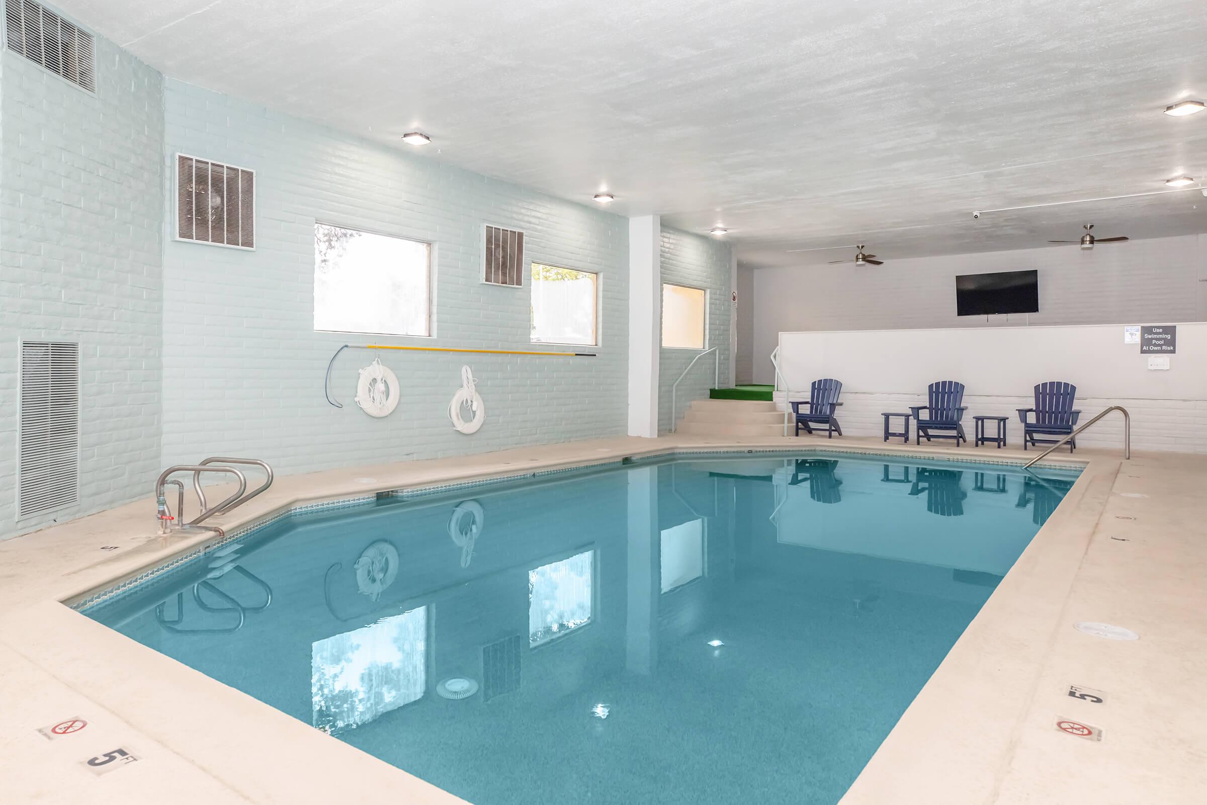Indoor swimming pool with clear blue water, light-colored walls, and a few lounge chairs nearby. The pool area is well-lit with natural light coming through windows. A small green area is visible in the background, and a television monitor is mounted on the wall. Safety equipment is also present.