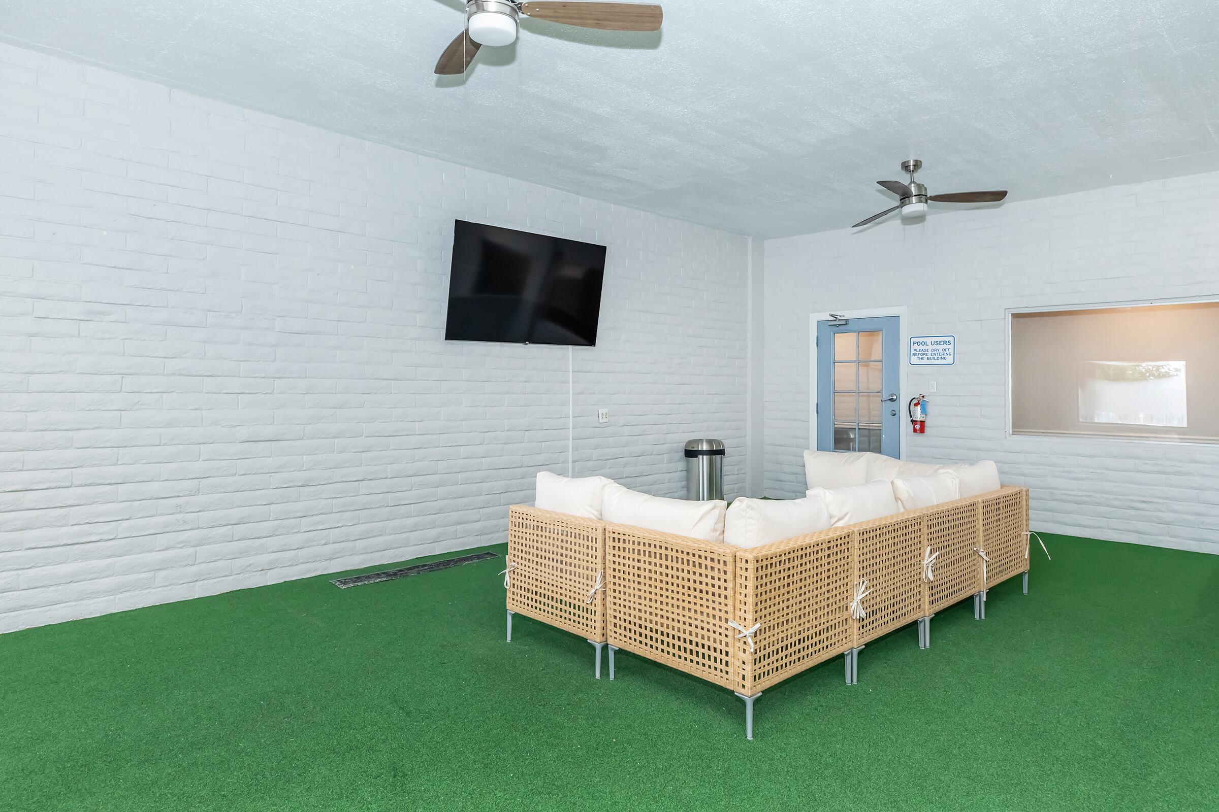 A cozy lounge area featuring a light-colored, wicker sofa with white cushions, set on green artificial grass. A flat-screen TV is mounted on the wall, and there are ceiling fans for ventilation. Windows allow natural light, creating a welcoming atmosphere for relaxation.