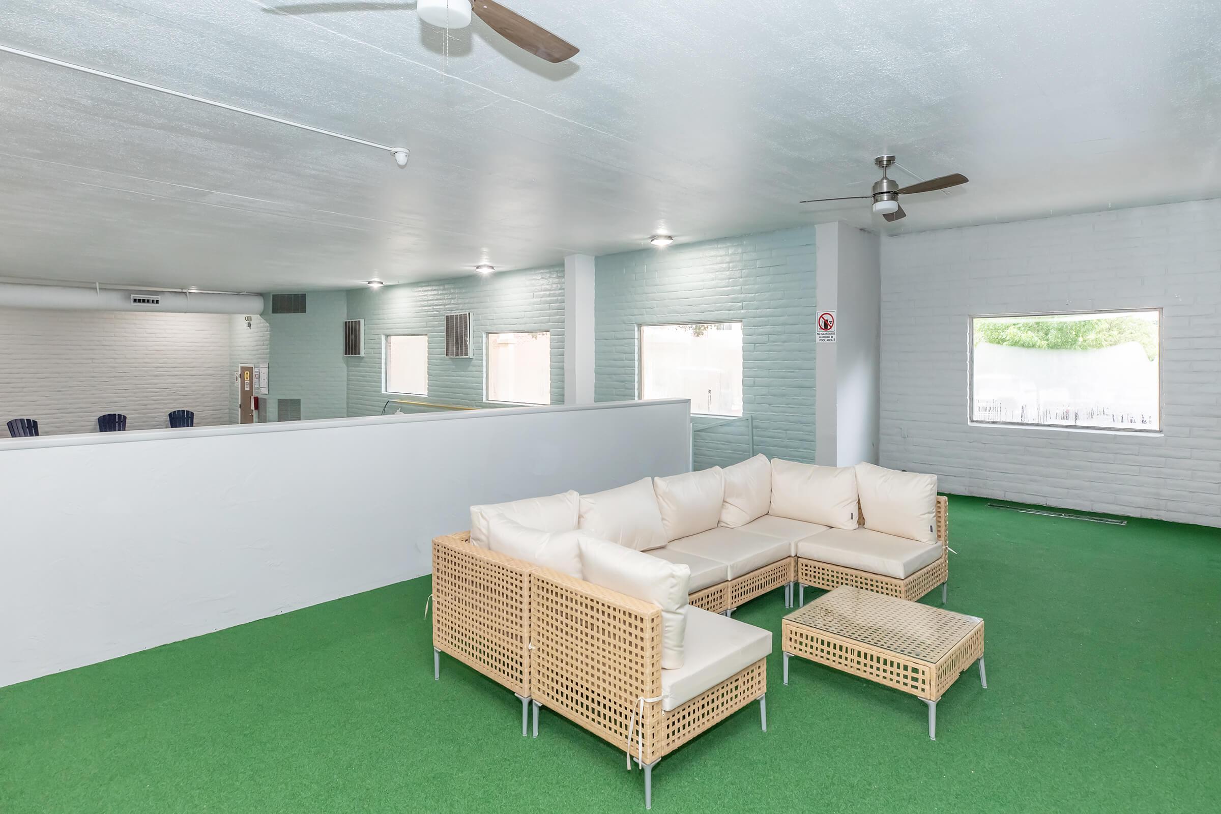 A spacious indoor area featuring a modern wicker sofa set with cushions, arranged around a small table. The floor is covered with green artificial grass, and there are several windows allowing natural light to fill the space, creating a bright and inviting atmosphere.