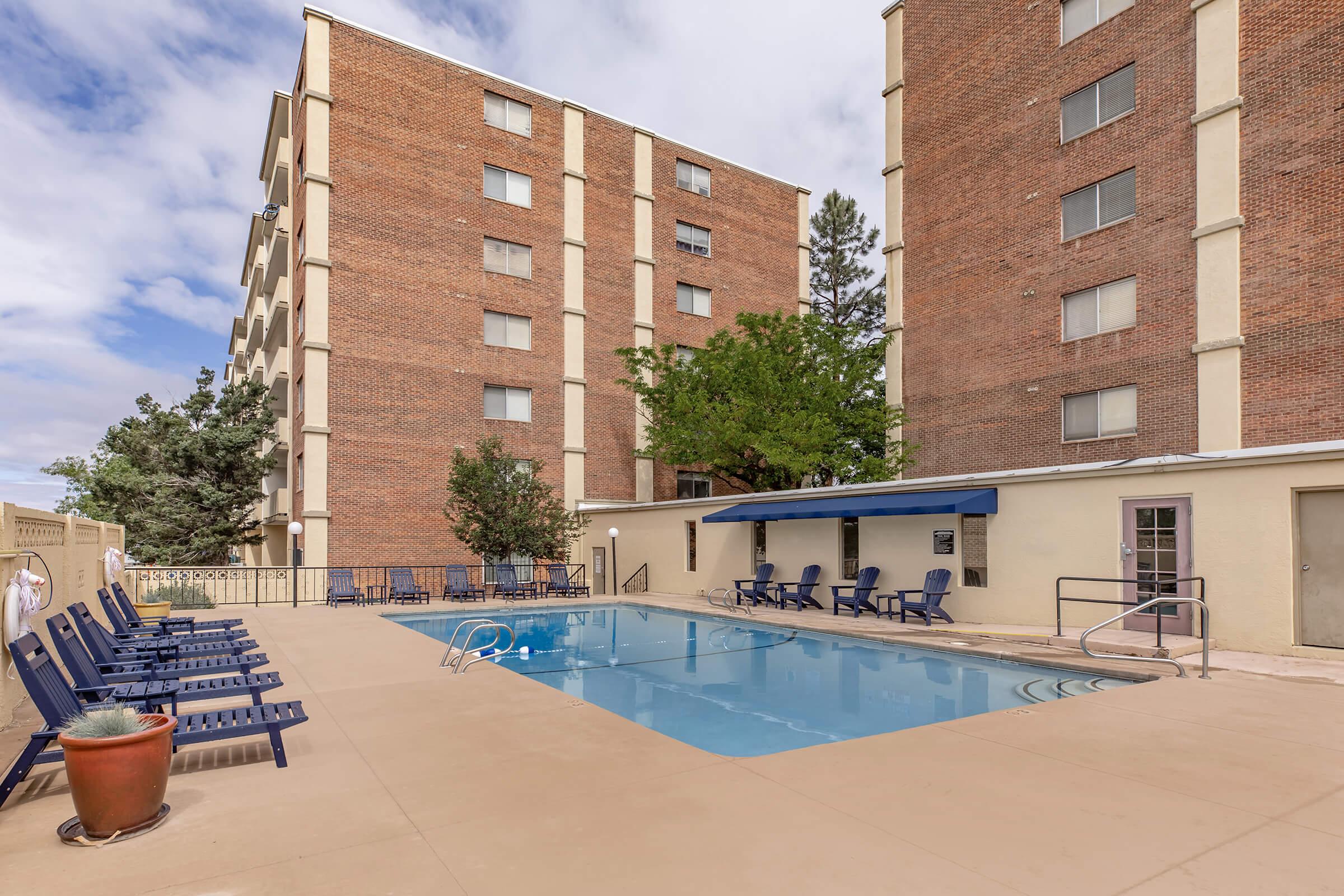 A swimming pool surrounded by lounge chairs, located in a courtyard of a brick apartment building. Lush greenery is visible nearby, and there is a shaded area with seating. The clouded sky adds a serene atmosphere to the outdoor space.