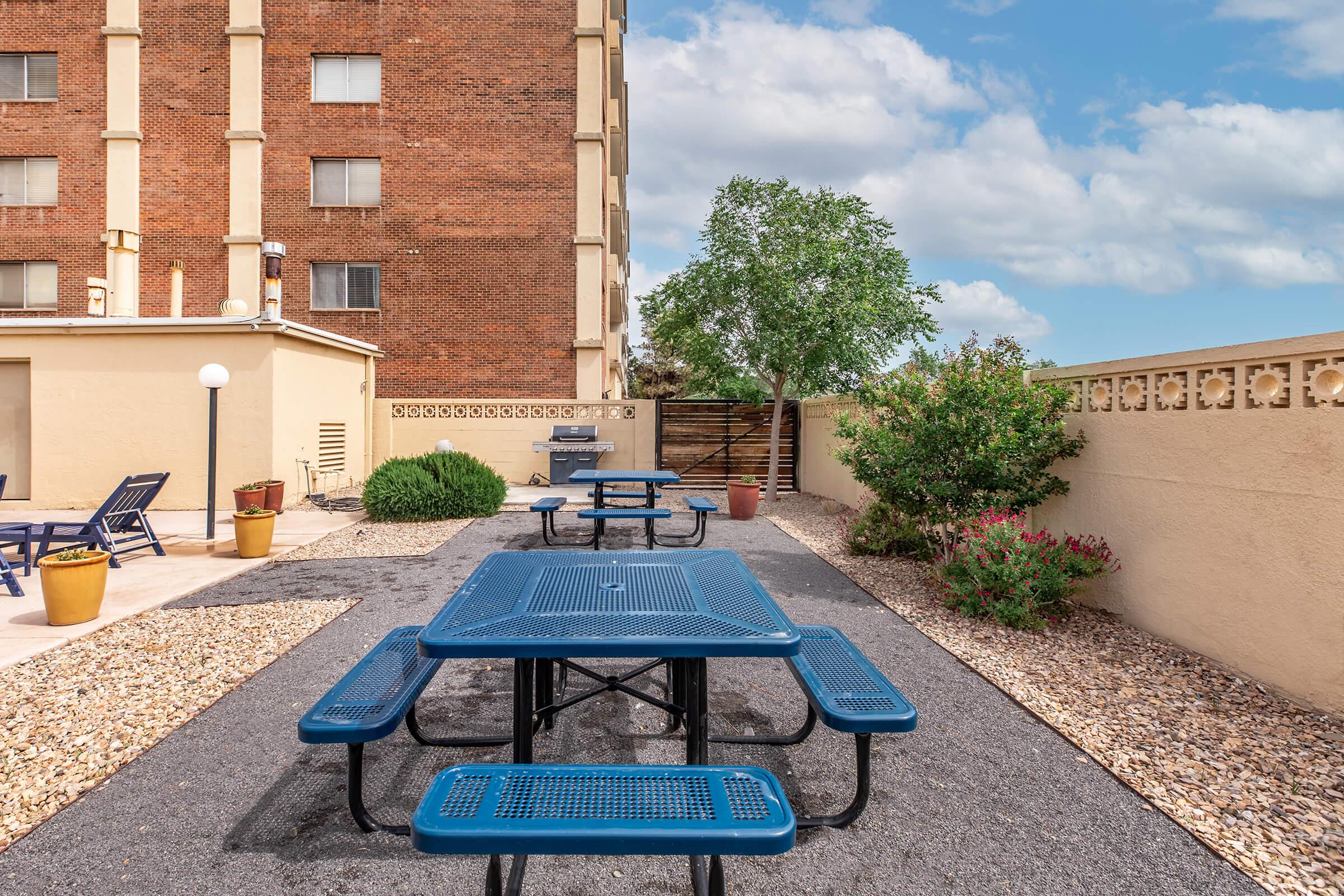 Outdoor recreational area featuring blue picnic tables surrounded by gravel, potted plants, and a wall. In the background, a multi-story brick building and trees can be seen under a partly cloudy sky.