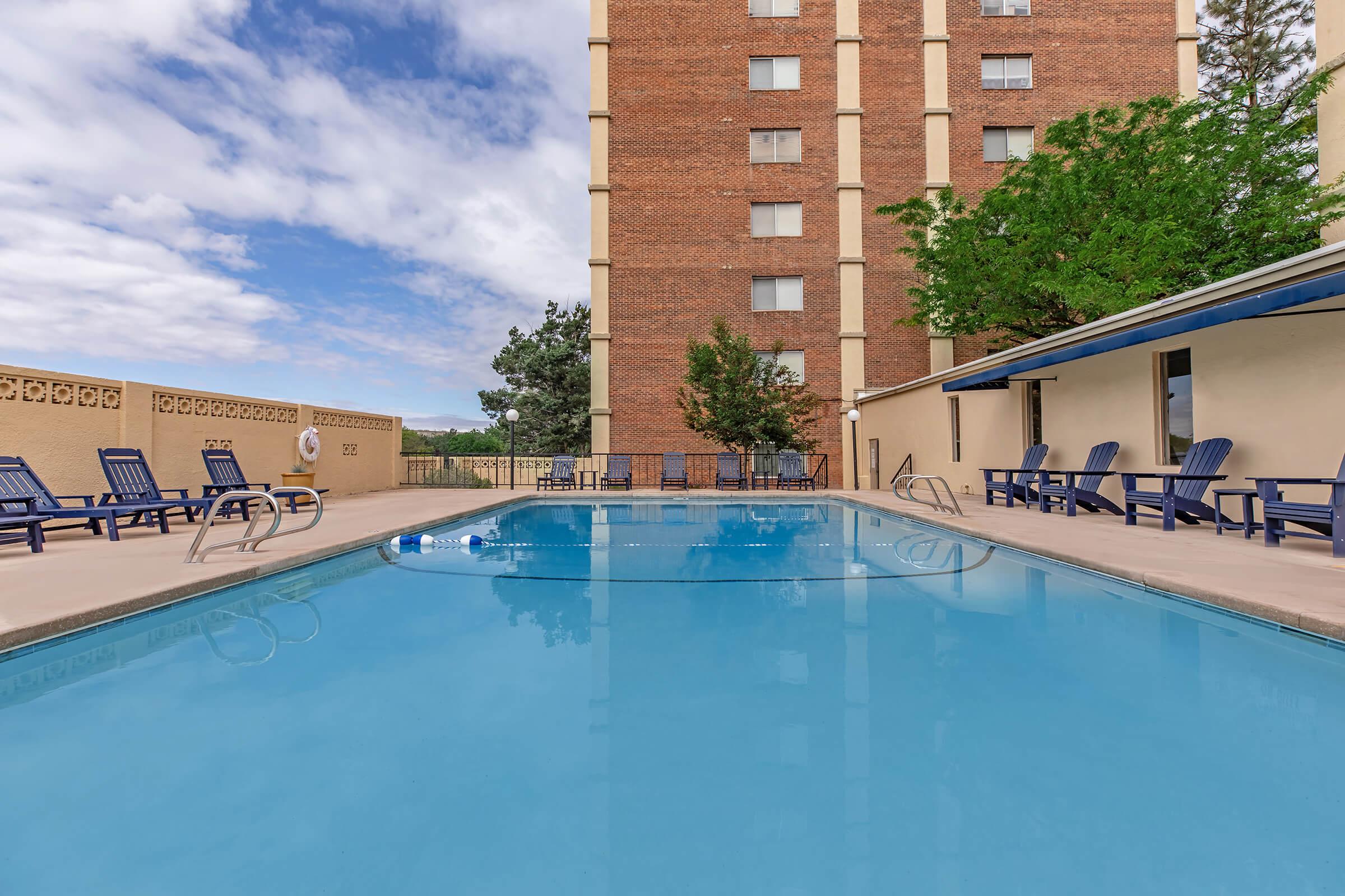 A clear blue swimming pool surrounded by lounge chairs, with a brick building in the background and trees nearby. The sky is partly cloudy, adding to the inviting atmosphere of the pool area.