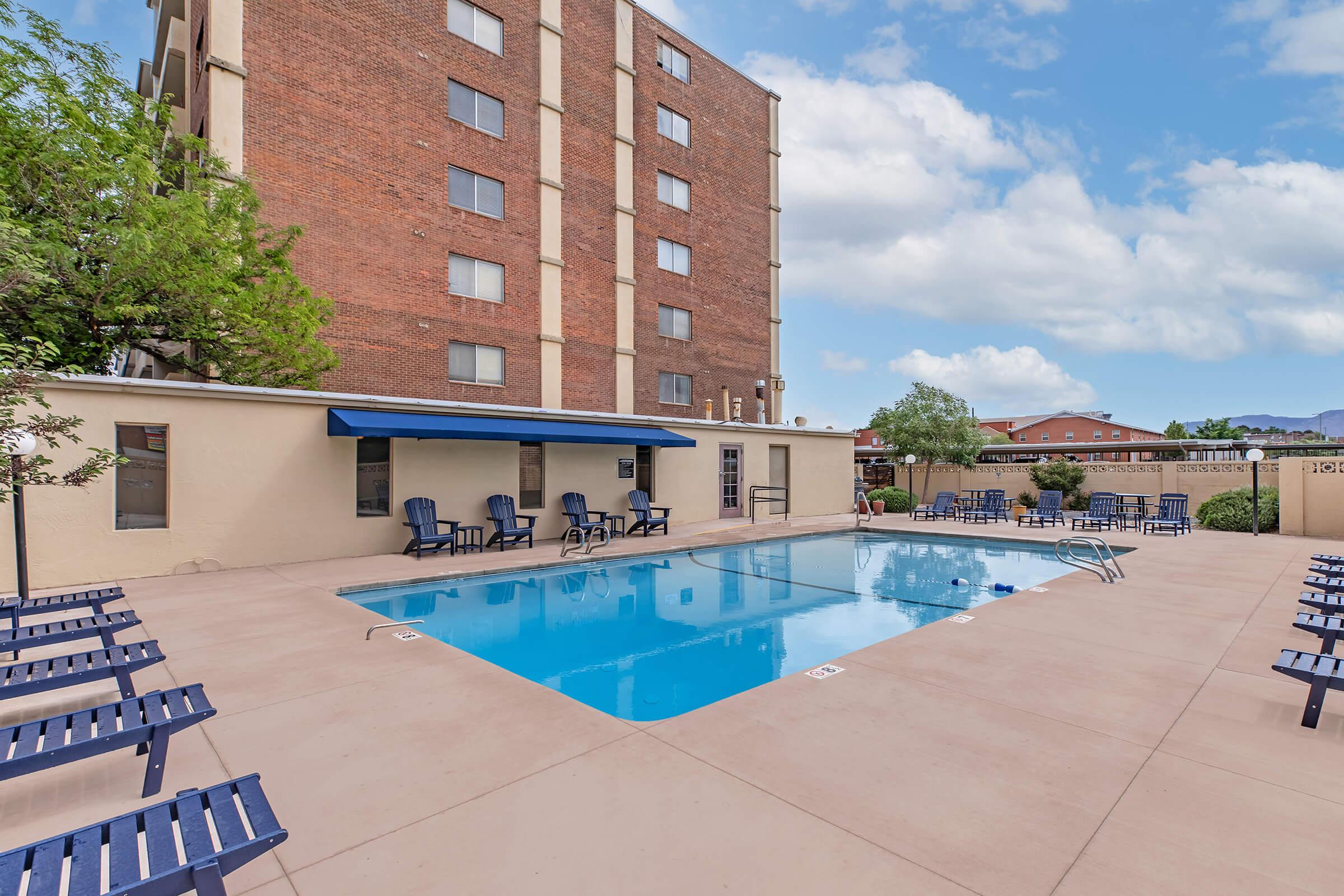 A clean outdoor swimming pool surrounded by lounge chairs, with a blue awning and a backdrop of a multi-story brick building and trees. The scene is bright and inviting, ideal for relaxation and leisure on a sunny day.