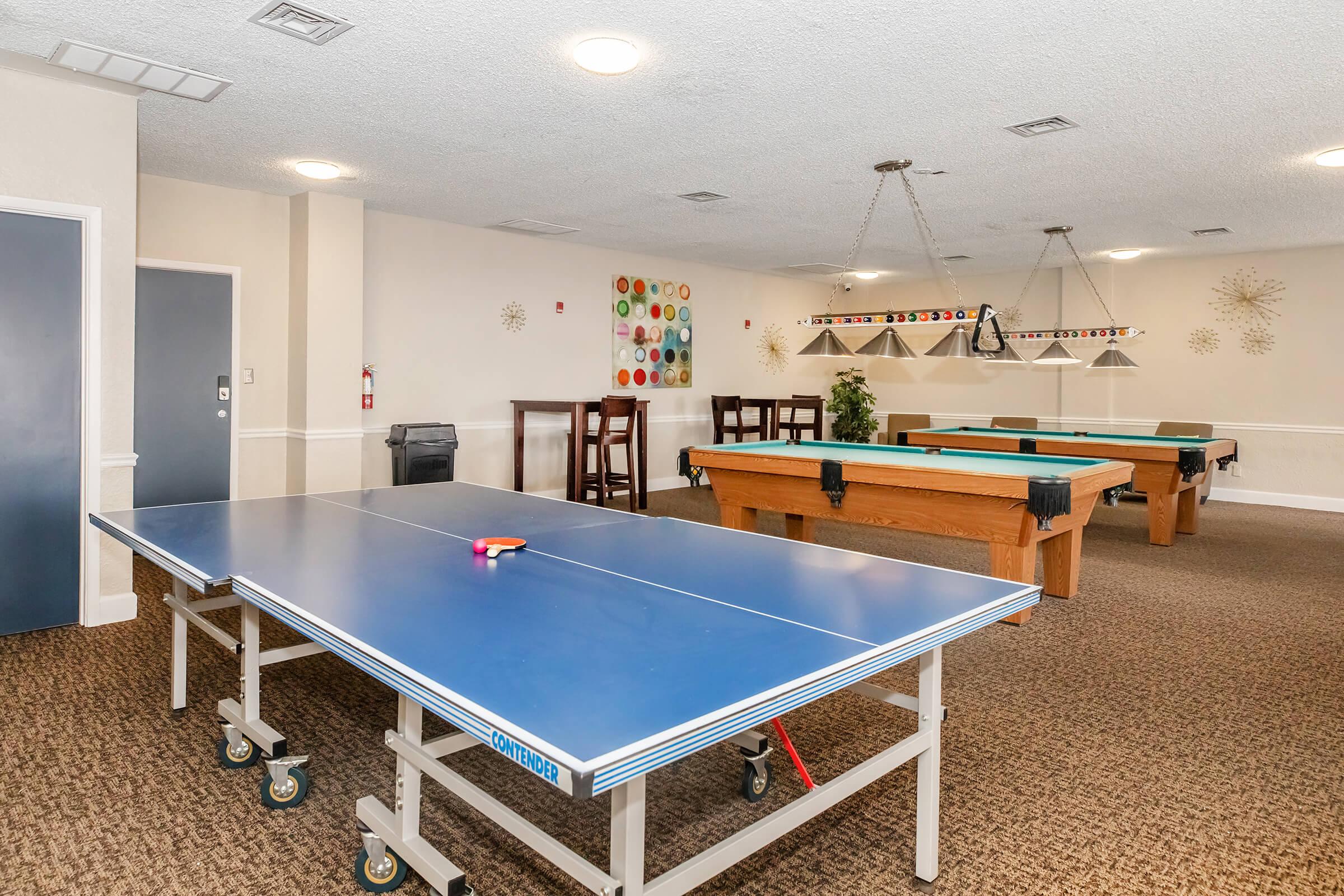 A recreational room featuring a blue ping pong table, several wooden bar stools, and two pool tables. The walls are adorned with colorful decorations, and there's a cozy carpeted floor with a warm lighting atmosphere.