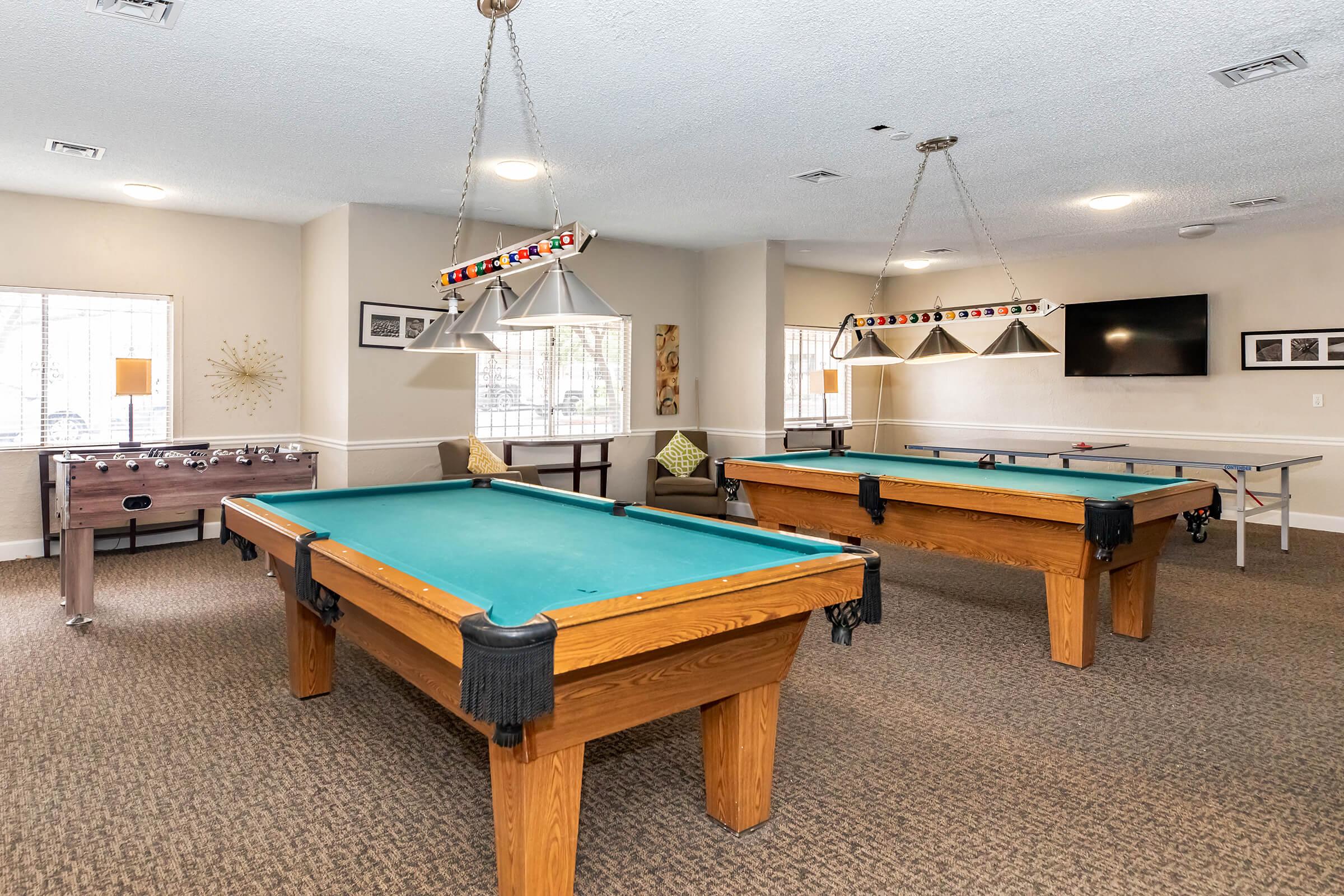 A recreational room featuring two pool tables with blue felt surfaces and wooden frames, a foosball table in the background, and a wall-mounted television. Soft lighting lamps hang above the tables, and there are comfortable seating areas with decorative pillows. The flooring is carpeted for a cozy atmosphere.