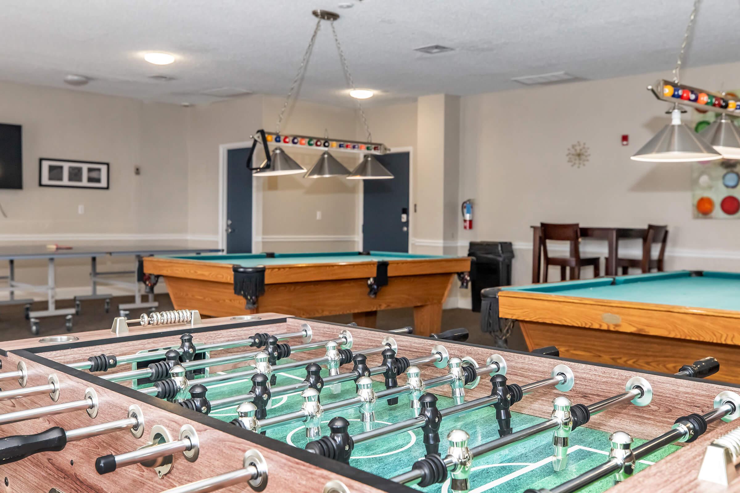 A game room featuring a foosball table in the foreground, with two pool tables visible in the background. The room is well-lit, with overhead lights and a casual setting that includes a seating area with chairs and a table. Walls are painted in neutral tones with minimal decor.