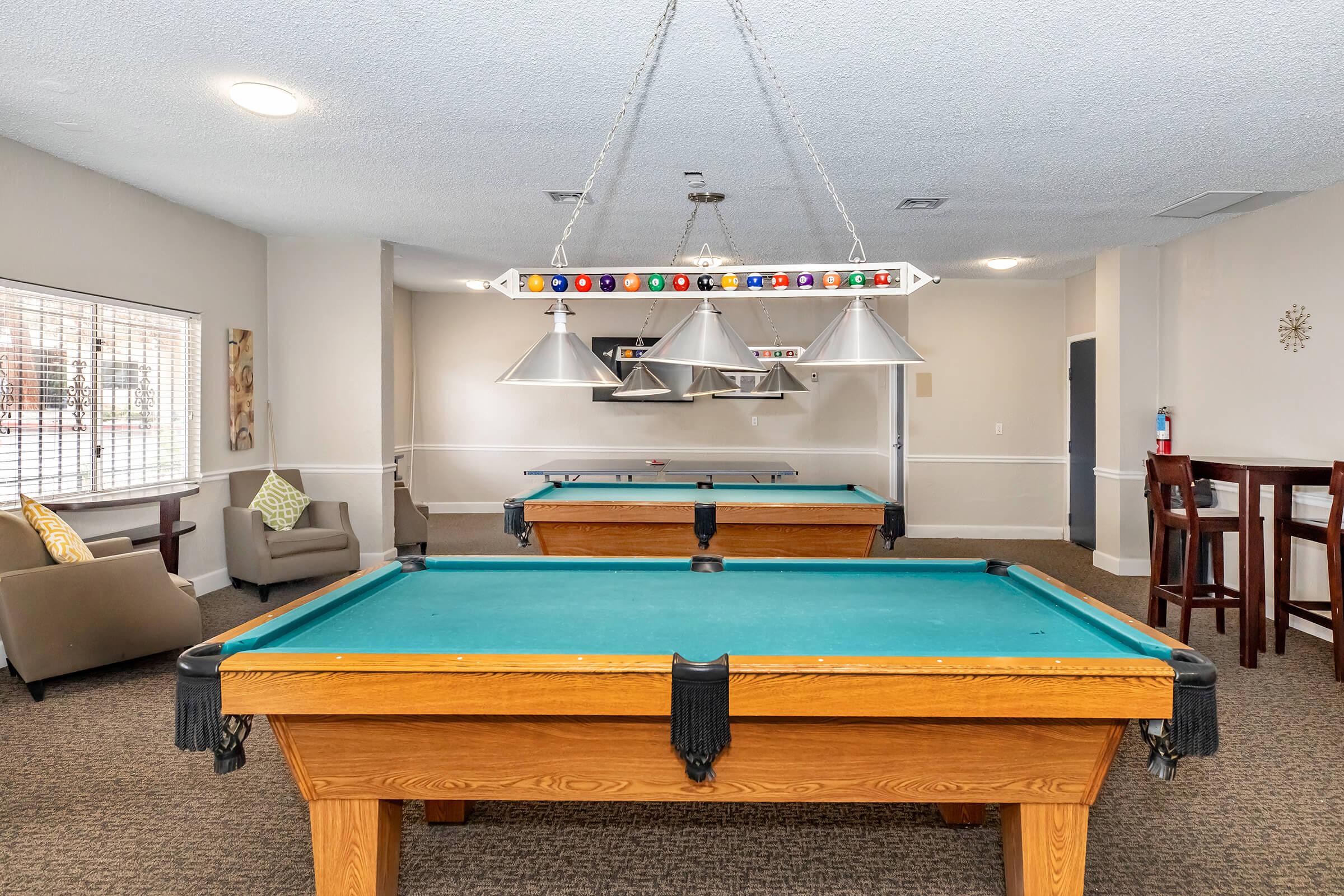 Two pool tables with green felt are set up in a well-lit recreational room. Above the tables, multiple pendant lights hang, providing illumination. The space features beige walls and carpet, with a seating area visible on one side, including a chair and decorative pillows. Natural light comes through a large window.