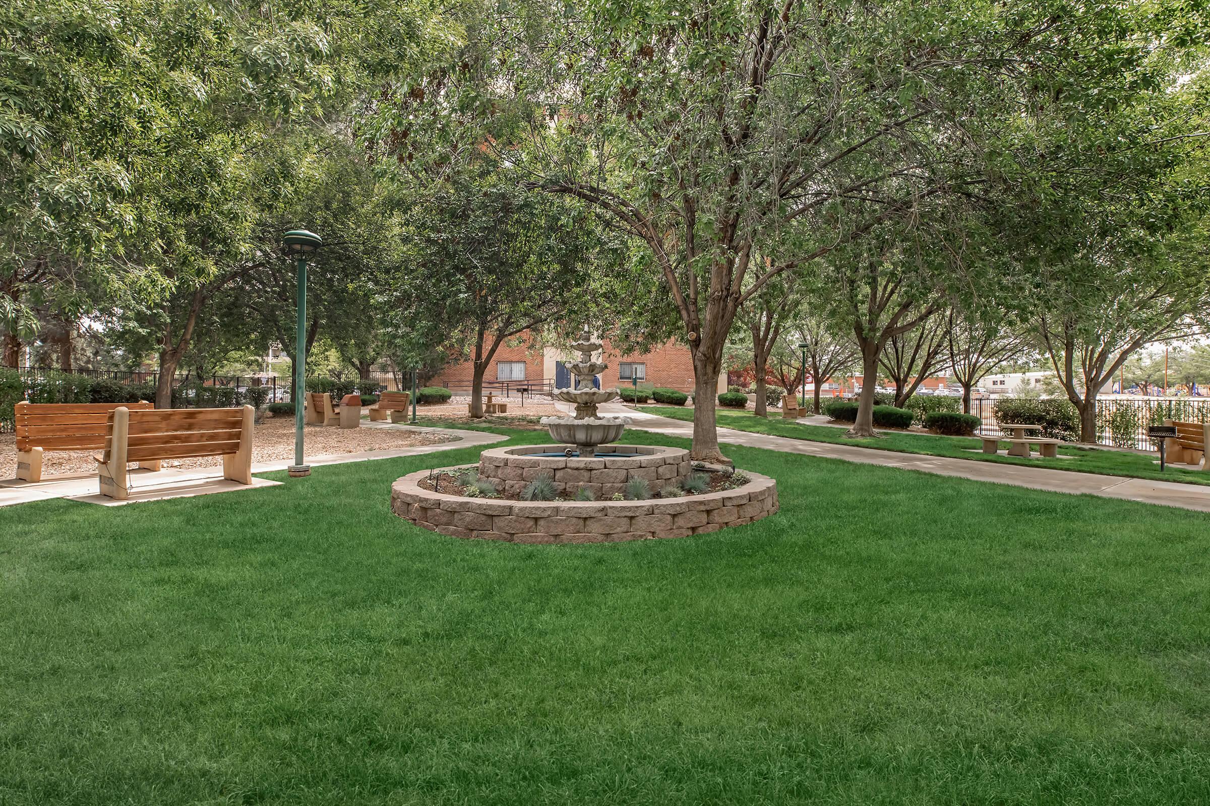 A green park with well-manicured grass, featuring a central stone fountain surrounded by neatly arranged flower beds. There are benches for seating under the shade of large trees, with pathways leading through the area, creating a peaceful and inviting outdoor space.