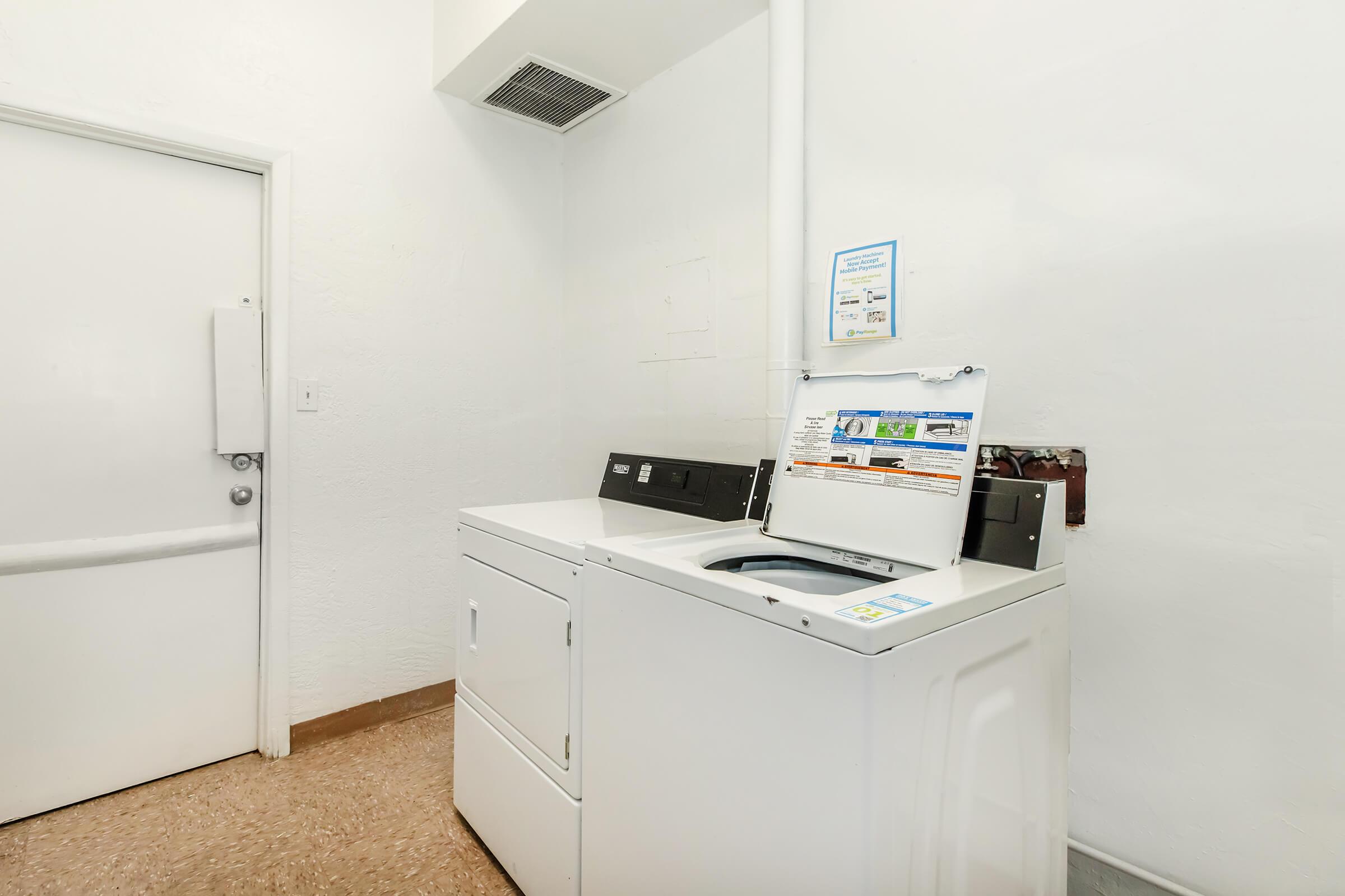 A clean, well-lit laundry room featuring a washing machine and dryer against a white wall. A door is visible on the left side, and a sign is posted above the washing machine, likely providing instructions or information about the appliances. The floor is covered in brown linoleum tiles.