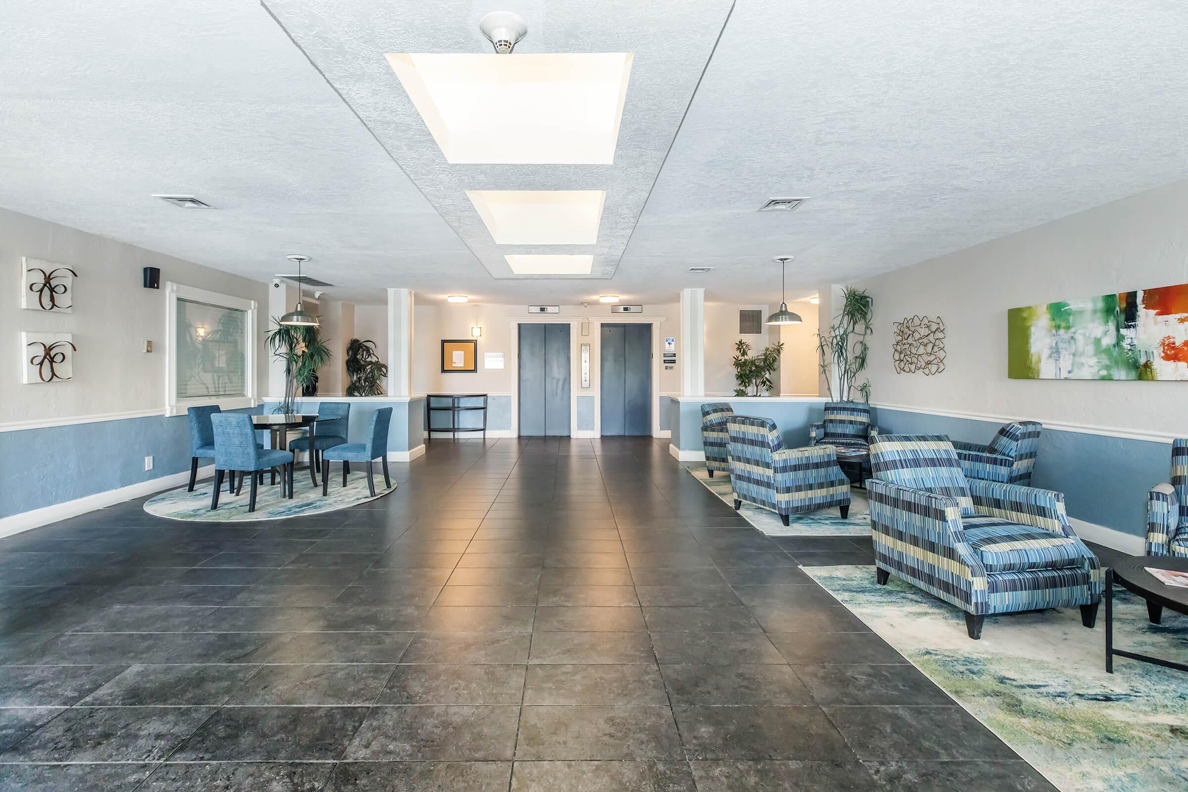 A spacious hotel lobby featuring modern seating arrangements, including two striped armchairs and matching rugs. There is a small round table with four blue chairs, and the area is well-lit with overhead lights. Potted plants are placed throughout, adding a touch of greenery to the decor.