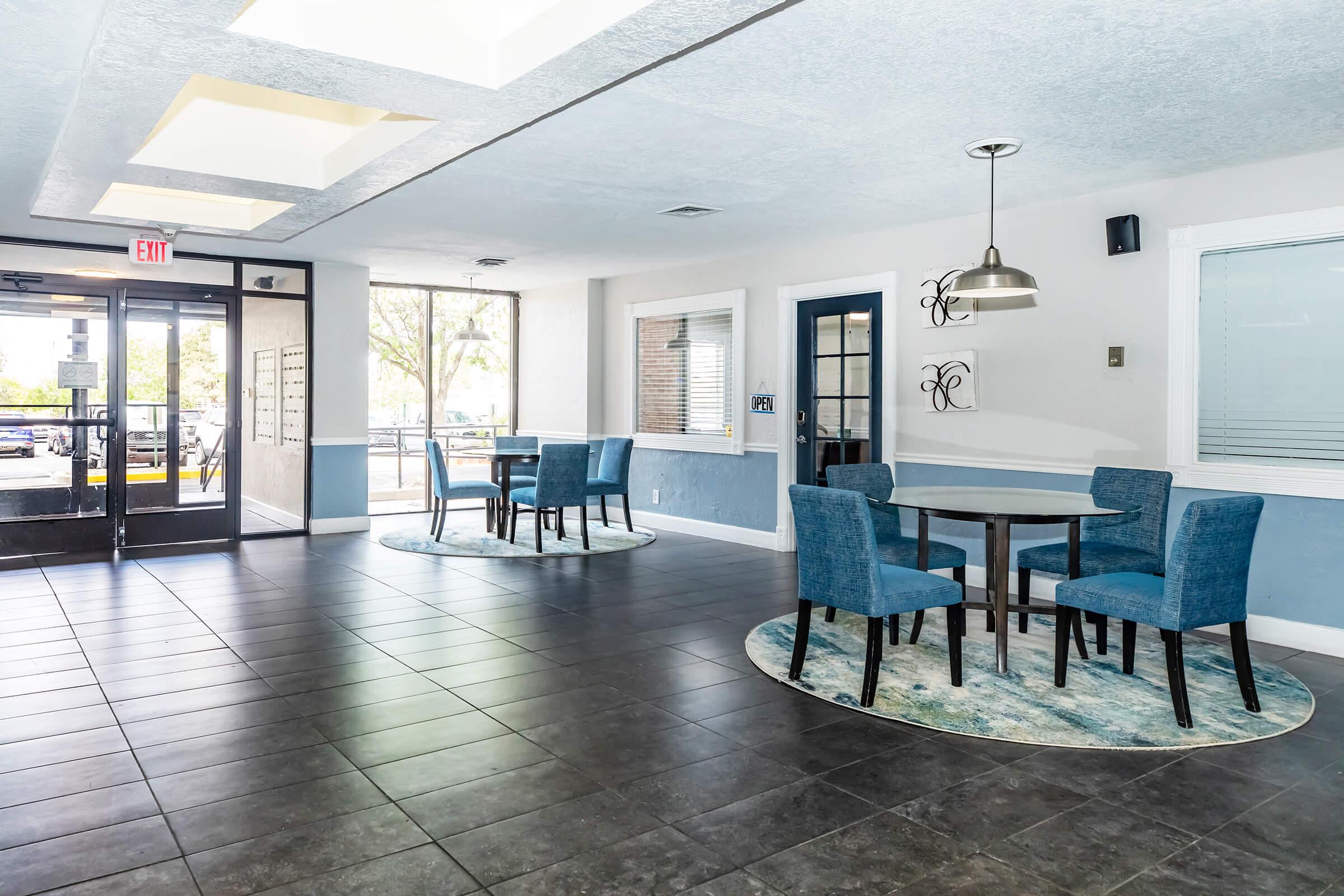 Bright and airy interior of a modern dining area featuring two tables with blue chairs on circular rugs. There are large windows allowing natural light, and a statement light fixture hanging from the ceiling. The walls are light-colored with accent details. The entrance is visible, leading to an outdoor space.