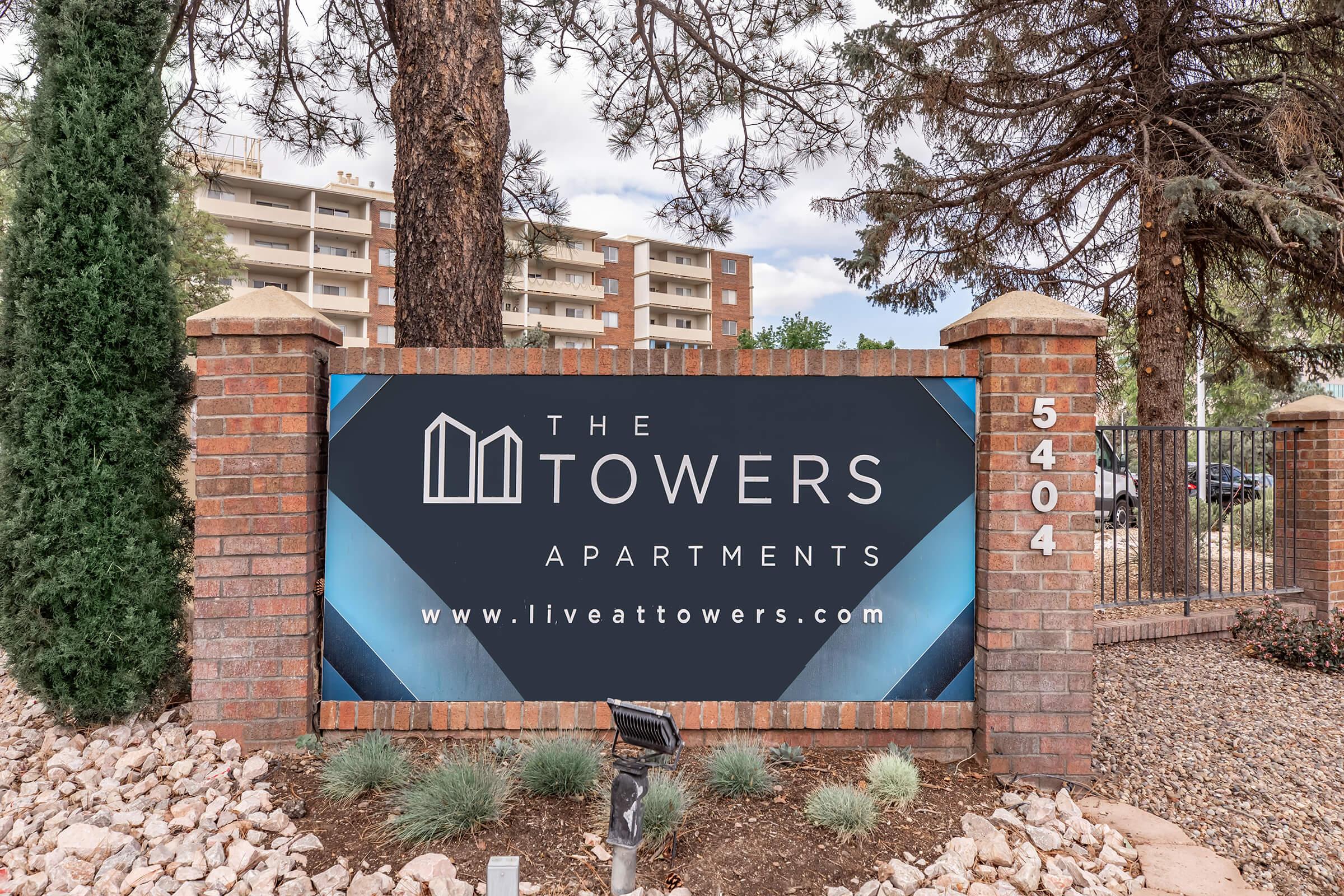 Sign for "The Towers Apartments" featuring a modern design with the website address www.liveattowers.com. The sign is set against a background of trees and landscaped rocks, indicating the location. The address shown is 5404, suggesting the site of the apartments.