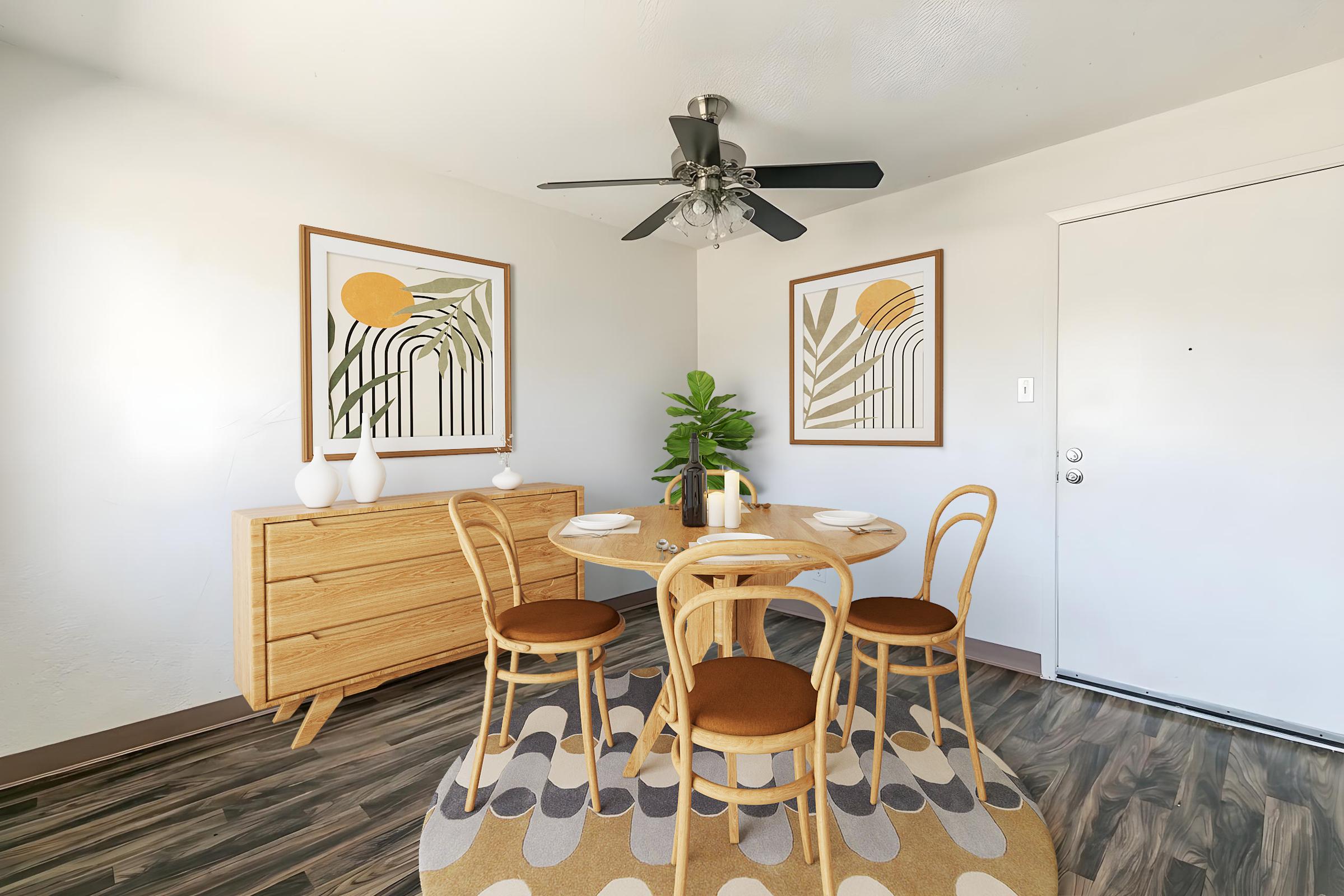 A cozy dining area featuring a round wooden table with four chairs, a decorative rug, and a light wood sideboard. Two framed artworks with abstract designs hang on the walls, and a potted plant adds greenery to the space. A ceiling fan is installed, and the entrance door is visible.