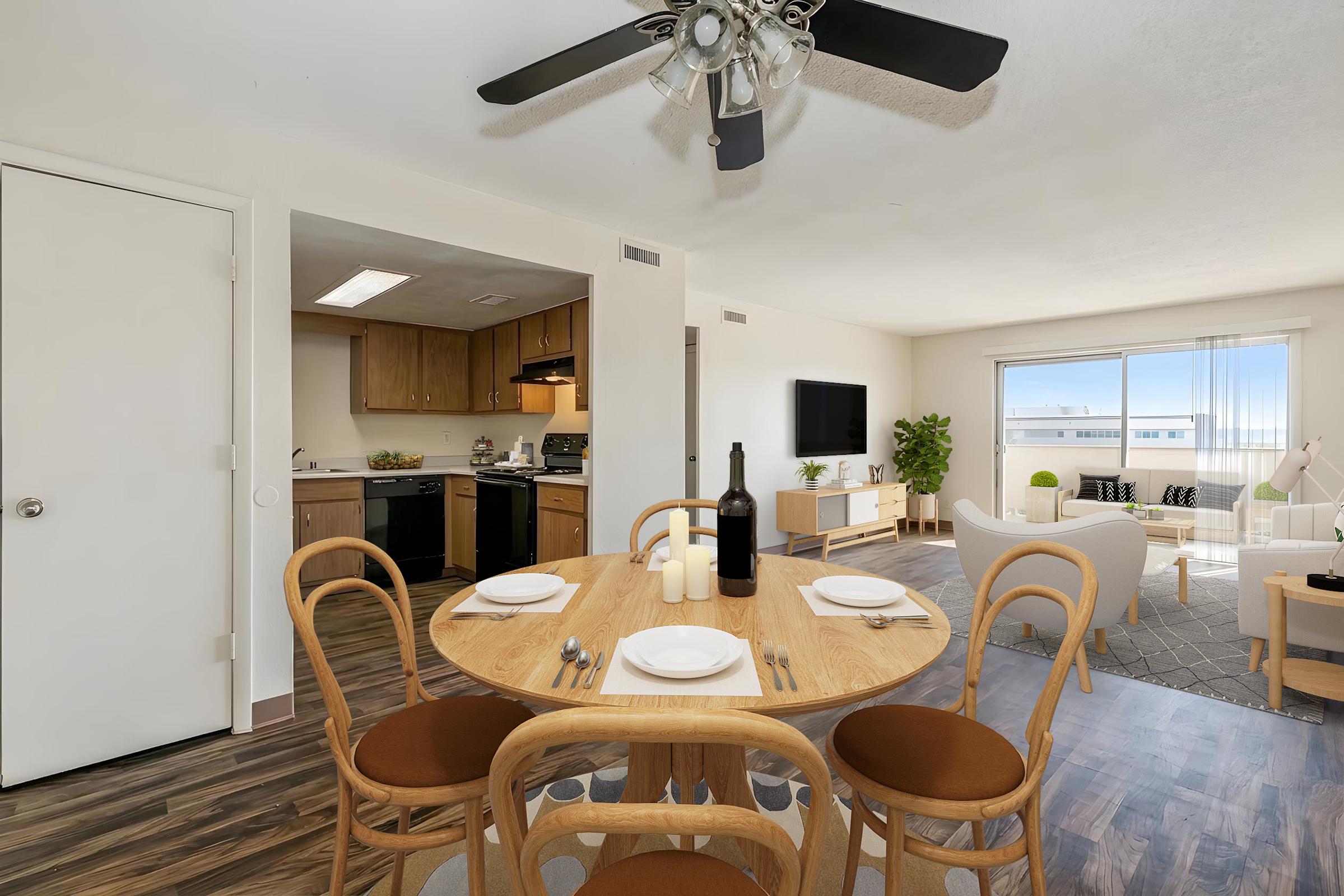 A modern dining area featuring a round wooden table set for four, with a wine bottle and candles. In the background, there’s a kitchen with wooden cabinets and a living room with a white sofa and large windows, creating a bright and inviting atmosphere. A ceiling fan is visible above the dining table.