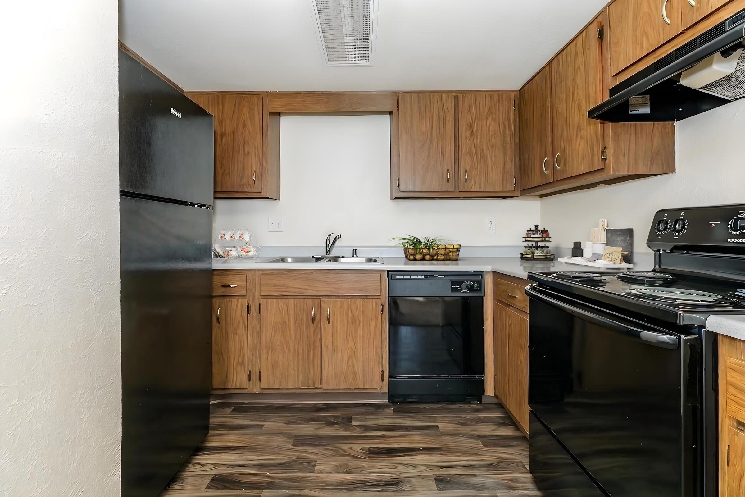 A modern kitchen featuring wooden cabinets, black appliances including a refrigerator, stove, and dishwasher. The countertop is light grey, with a sink under a window. Decor includes a small plant and a bowl of fruit on the counter. The flooring is a dark wood style, creating a warm, inviting atmosphere.