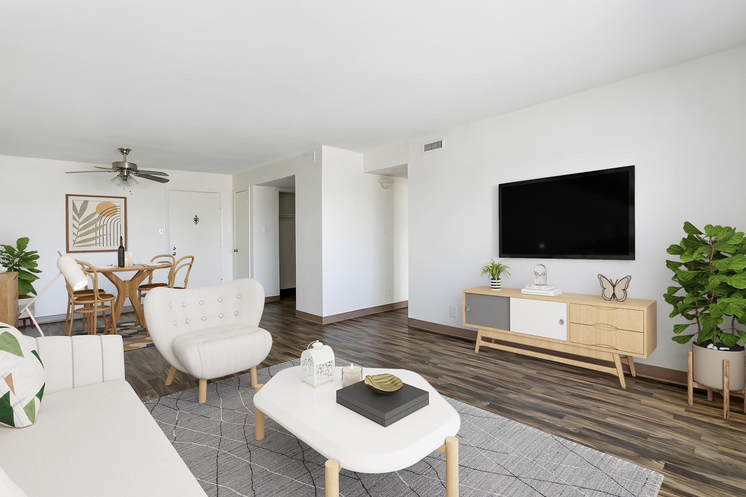 A modern living room featuring a light-colored sofa with decorative pillows, a white coffee table with a black tray, and a flat-screen TV mounted on a wooden cabinet. There are green plants in the corners, a dining area with a round table and chairs in the background, and light-colored walls and flooring.