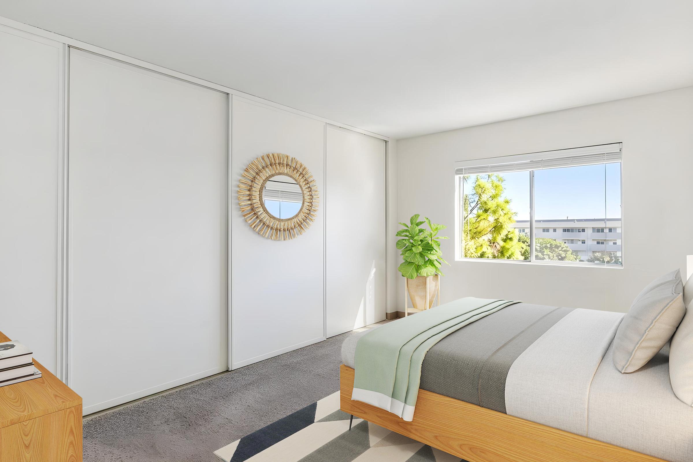 A modern bedroom featuring a bed with gray and white bedding, a decorative round mirror on the wall, a window with natural light, and a potted plant near the window. The walls are white, and there's a wooden nightstand next to the bed, contributing to a minimalist aesthetic.