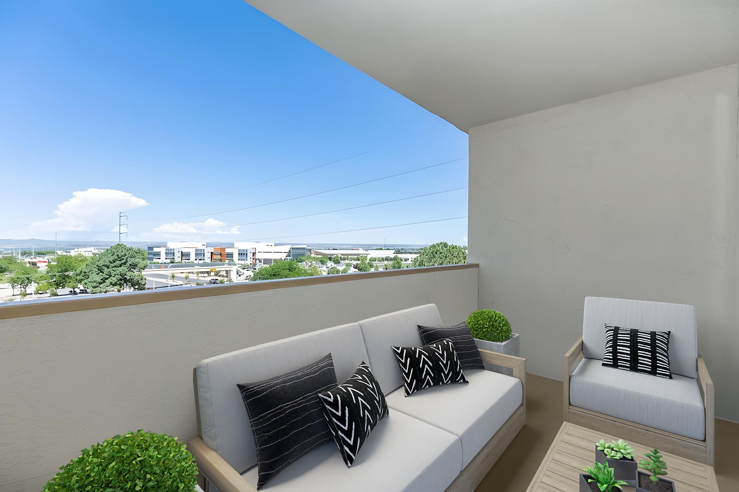 A modern balcony featuring a cozy seating area with light-colored furniture and decorative pillows. Potted plants are placed nearby, and in the background, a clear blue sky and urban landscape are visible, showcasing buildings and greenery.