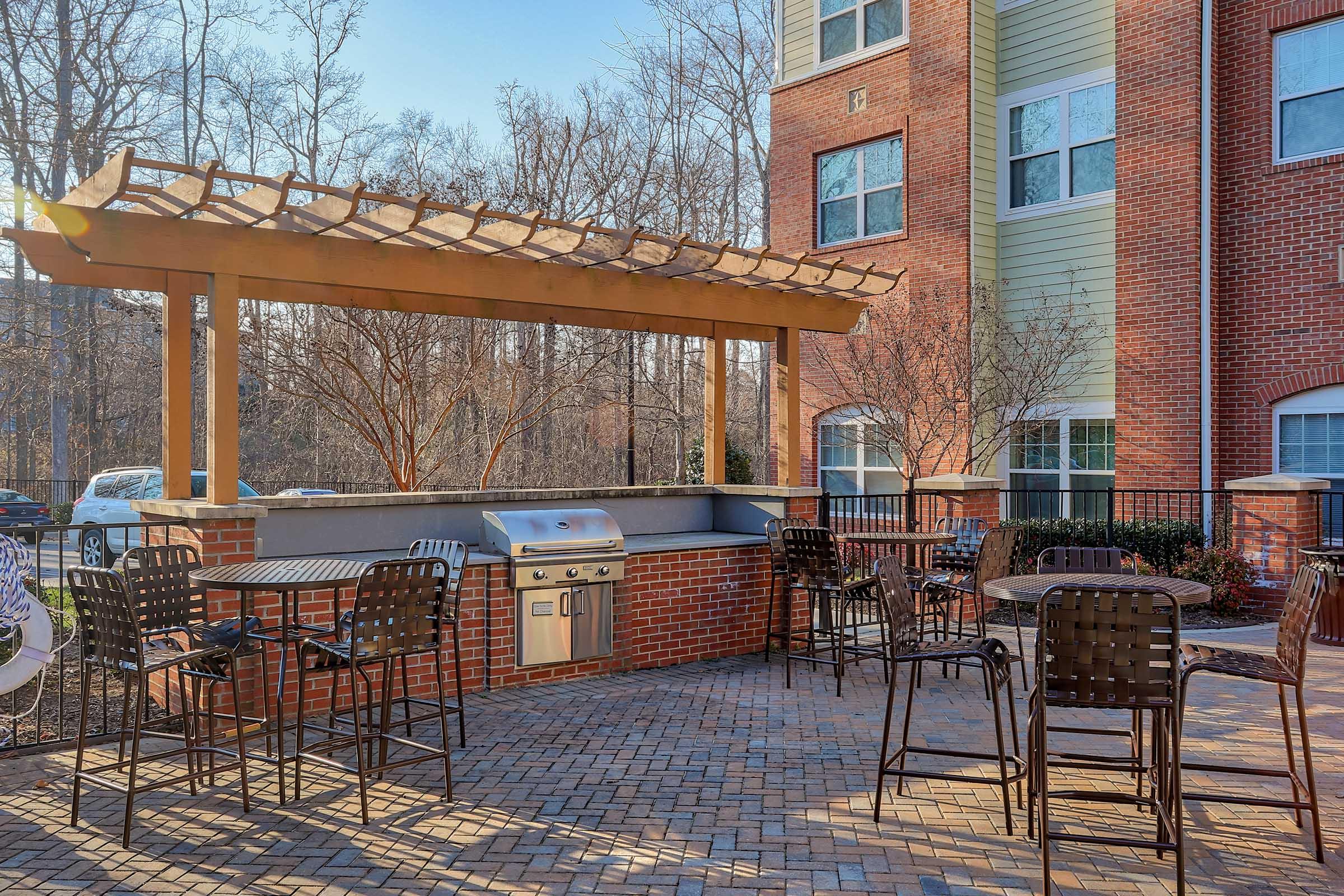 Outdoor patio area featuring a pergola with a grill, surrounded by bar stools and potted plants. The scene is set against a backdrop of a brick apartment building with large windows and trees in the background, under clear blue skies.