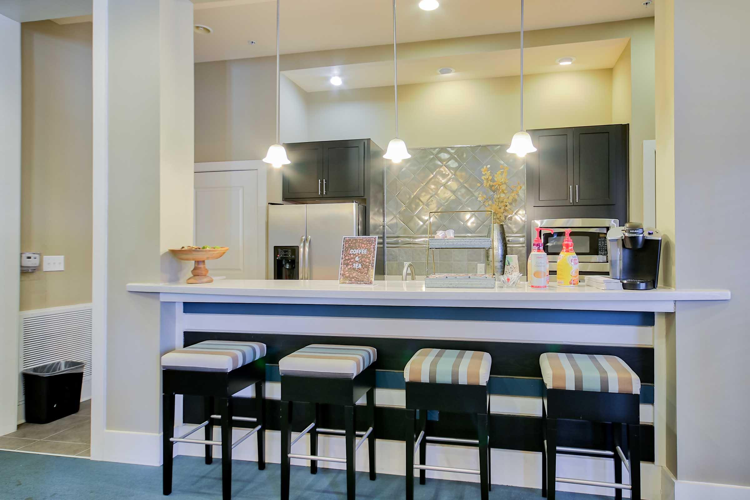 Modern kitchen interior featuring a bar counter with four striped stools. The space includes dark cabinetry with a tiled backsplash, a sink, and a coffee maker. Soft lighting fixtures hang above the counter, and a fruit bowl is placed on the surface. A trash bin is visible in the corner.