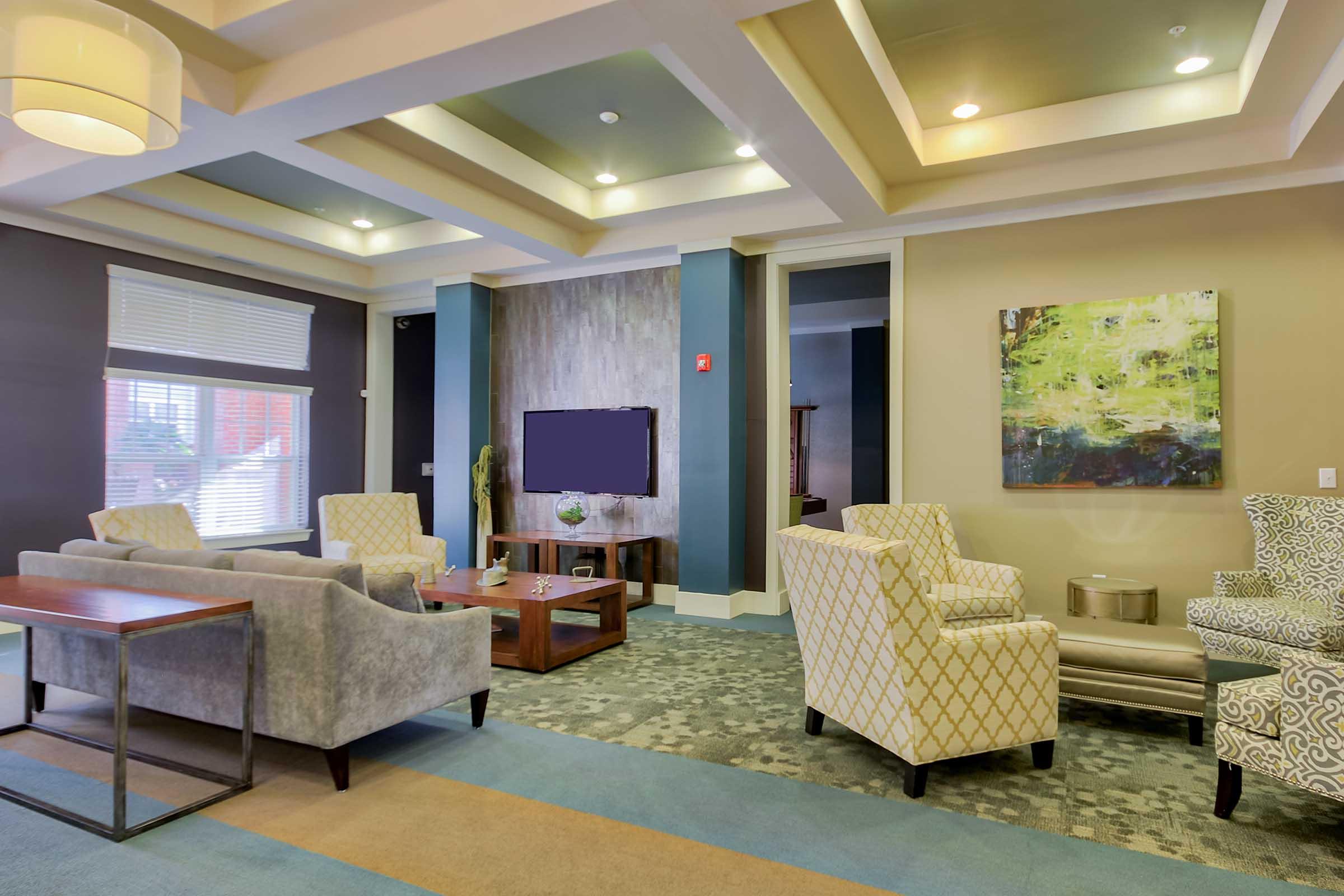 A cozy lounge area featuring gray and patterned armchairs, a wooden coffee table, and a flat-screen TV mounted on the wall. The room has soft lighting, modern decor, and a colorful abstract painting, with large windows allowing natural light to enter. The floor has a patterned carpet in shades of blue and beige.